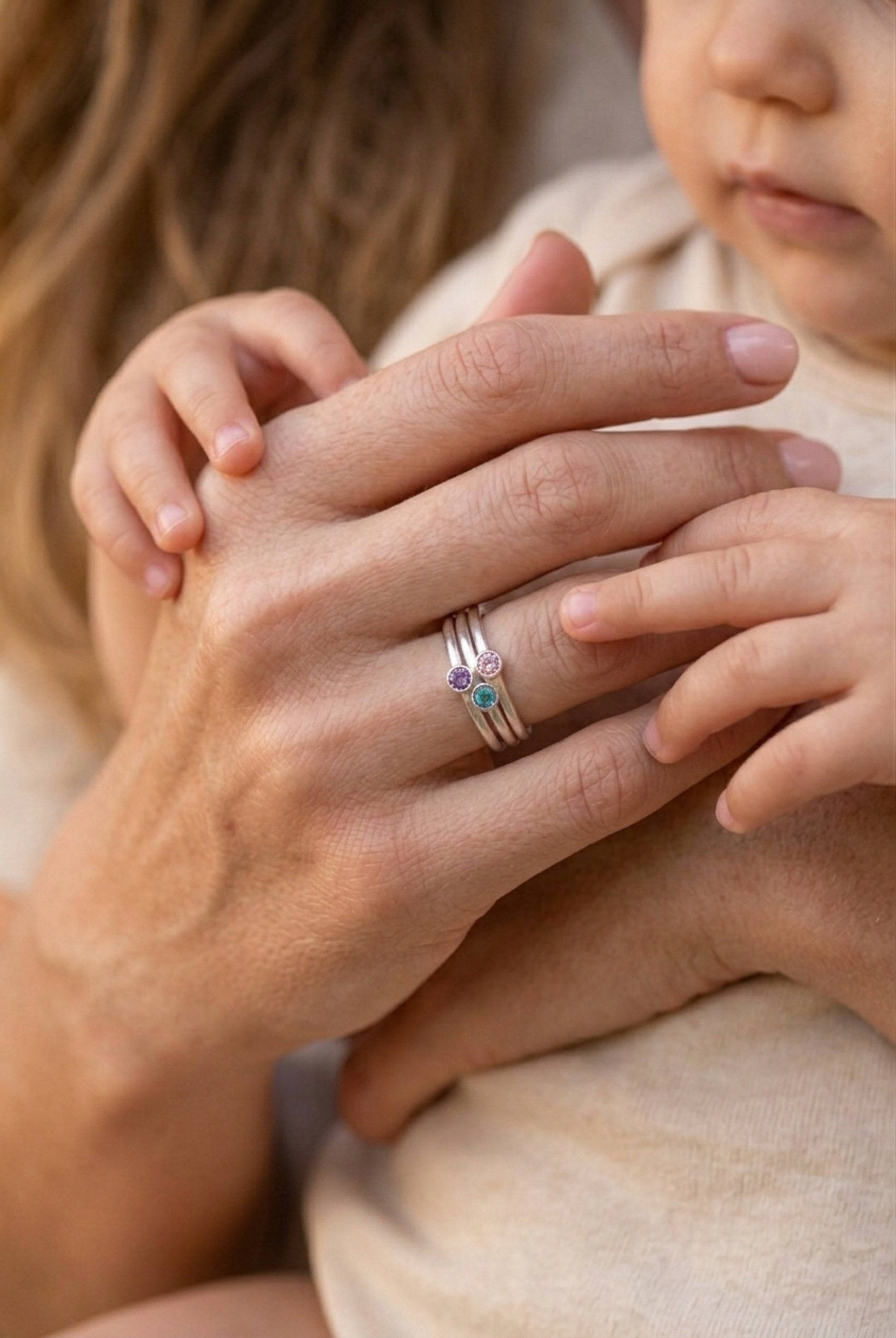 heartwarming close-up of an adult and baby's hands. The adult's right hand, with a polished light pink manicure, is in the foreground, wearing the stacked three-stone birthstone ring set (teal, pink, purple gems) from . Her hand is being tenderly held and explored by the soft, chubby hand of a baby. A partial view of the adult's blonde hair is on the left, and the baby's cheek is softly in focus on the right. The lighting is warm and natural.