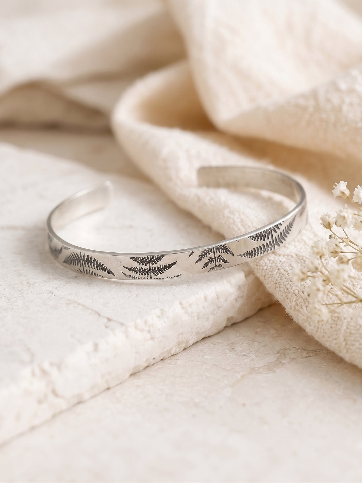 A sterling silver cuff bracelet with engraved fern leaf patterns, resting on a textured cream stone slab next to a soft beige fabric.