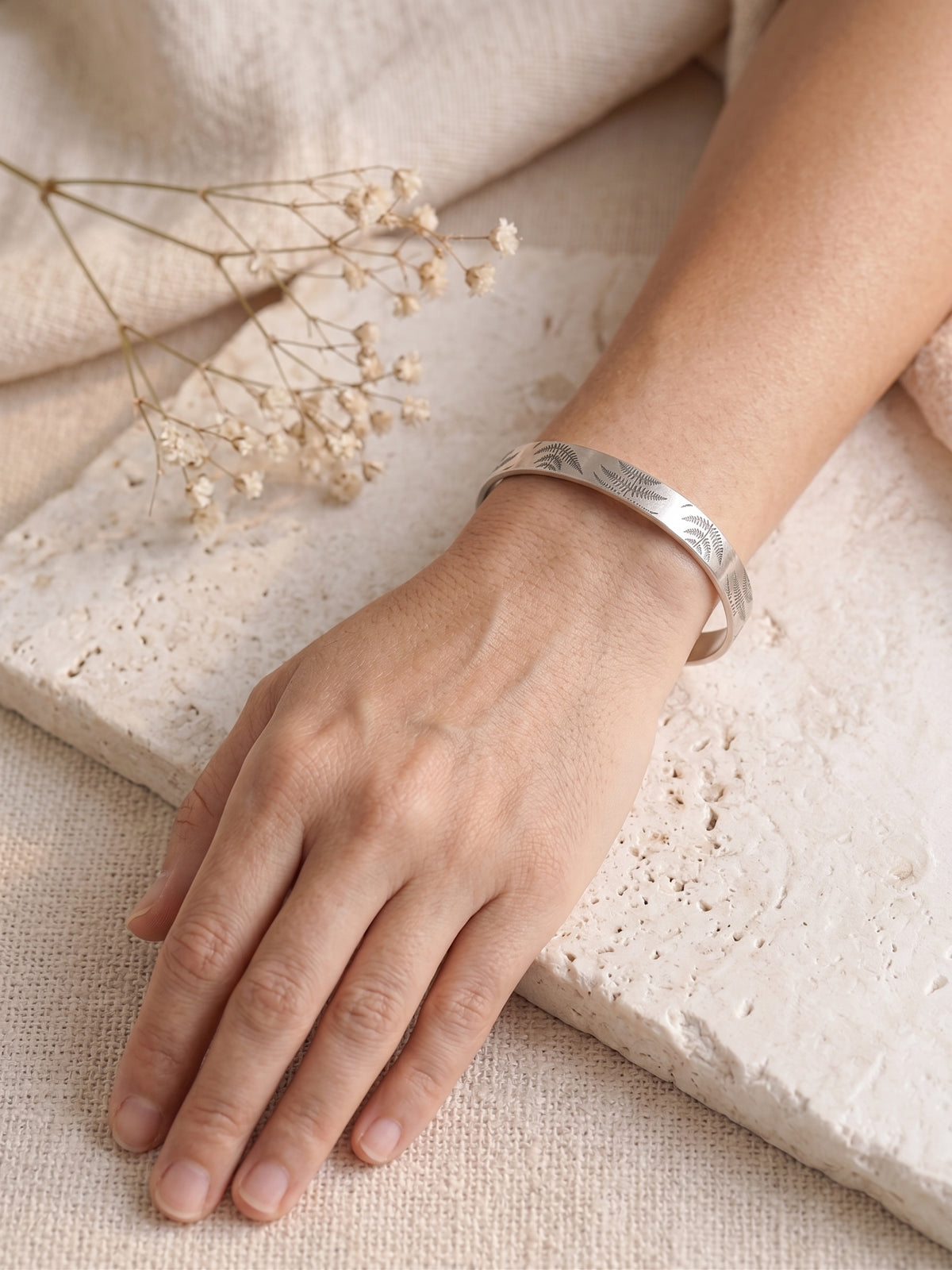 A sterling silvers metal cuff bracelet with a delicate fern pattern, worn on a person's wrist resting on a textured stone slab with dried baby's breath flowers.