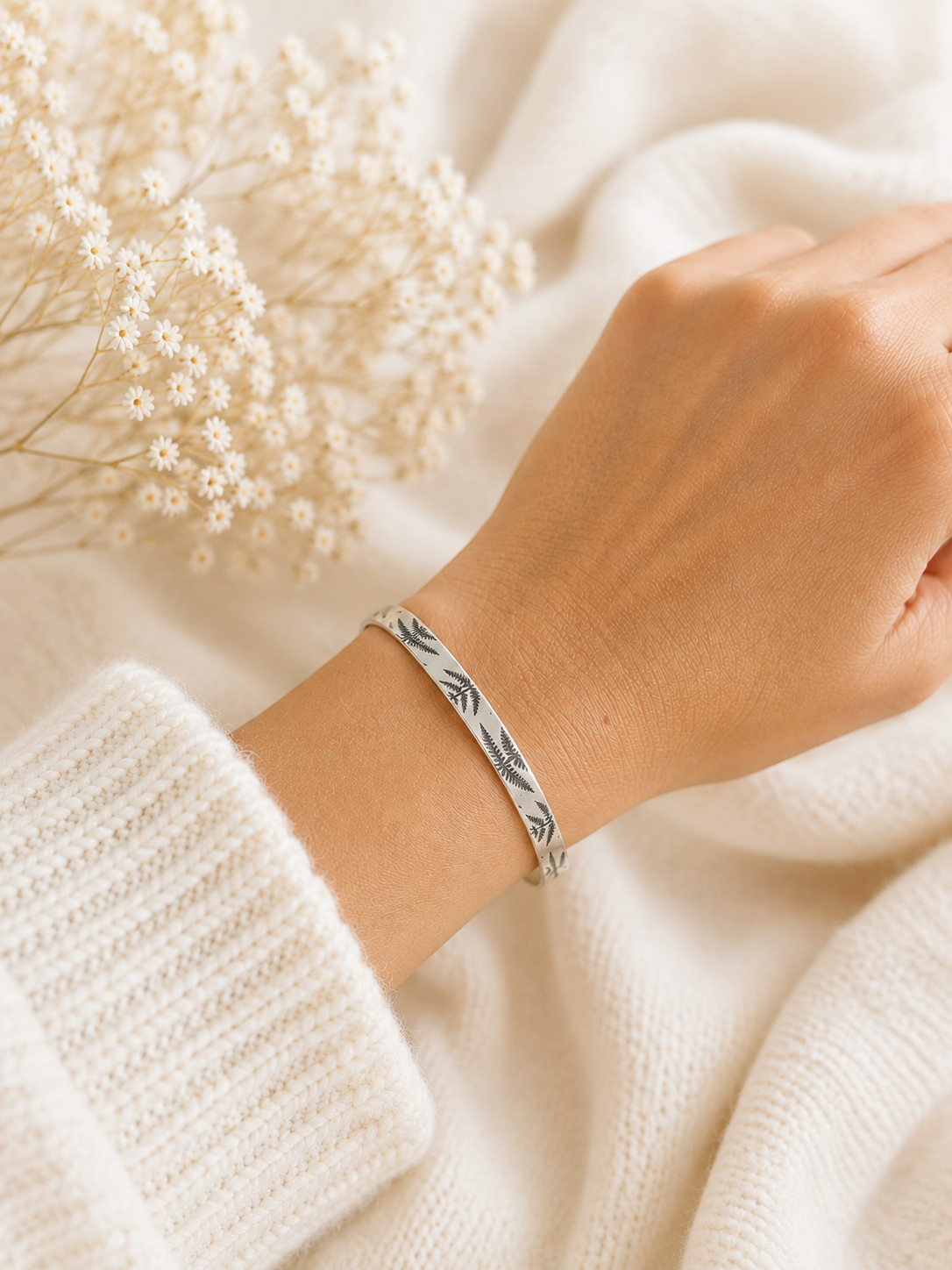 A silver-toned bangle bracelet with a fern leaf pattern, worn on a person's wrist over a white knit sweater, with dried white flowers in the background.