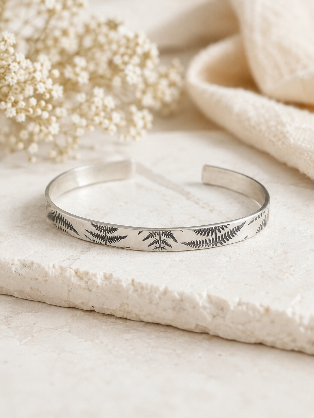A polished silver cuff bracelet with engraved fern leaf patterns, resting on a textured stone slab with dried flowers and a soft cream fabric in the background.