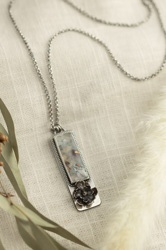 Sterling silver necklace with a sugar water flower agate pendant and succulent accent, laid flat on a beige fabric with dried eucalyptus leaves.
