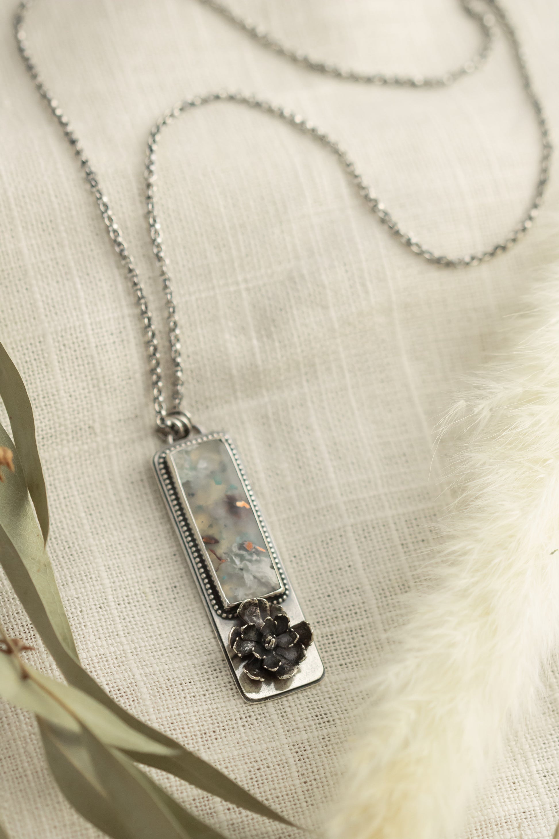 Sterling silver necklace with a sugar water flower agate pendant and succulent accent, laid flat on a beige fabric with dried eucalyptus leaves.
