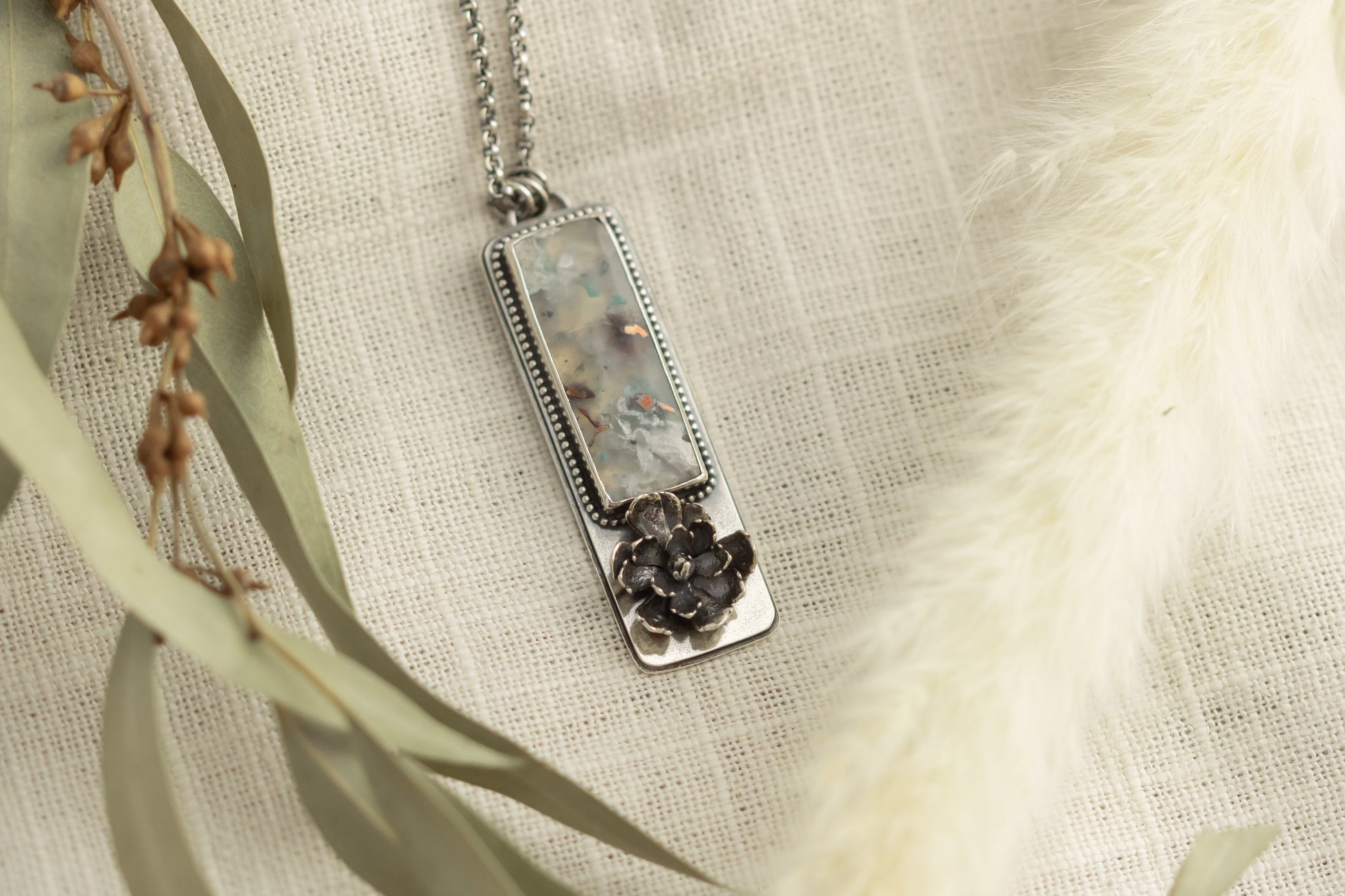 Sterling silver necklace with a sugar water flower agate pendant and succulent accent, laid flat on a beige fabric with dried eucalyptus leaves.

