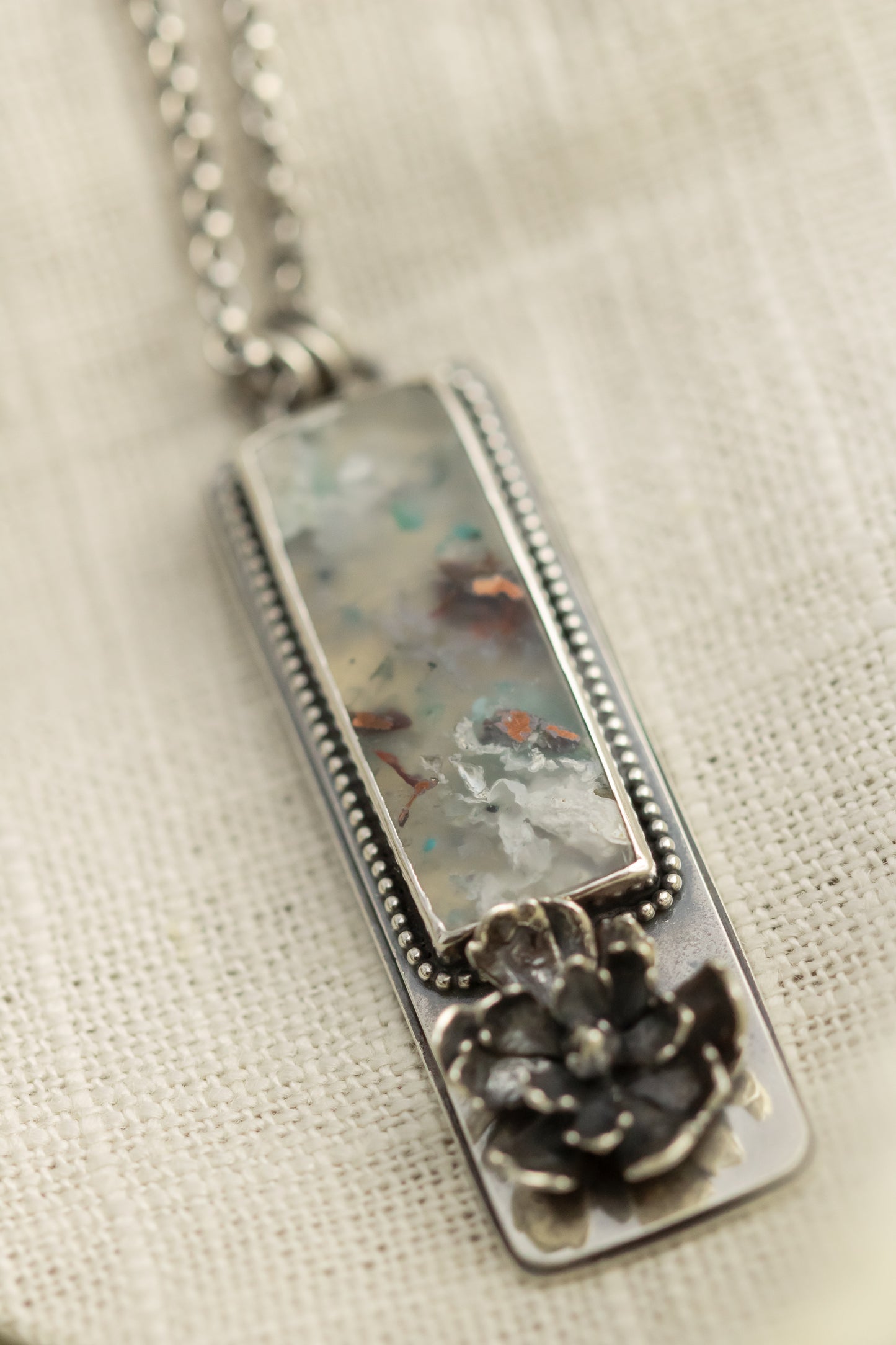 Sterling silver necklace with a sugar water flower agate pendant and succulent accent, laid flat on a beige fabric with dried eucalyptus leaves.
