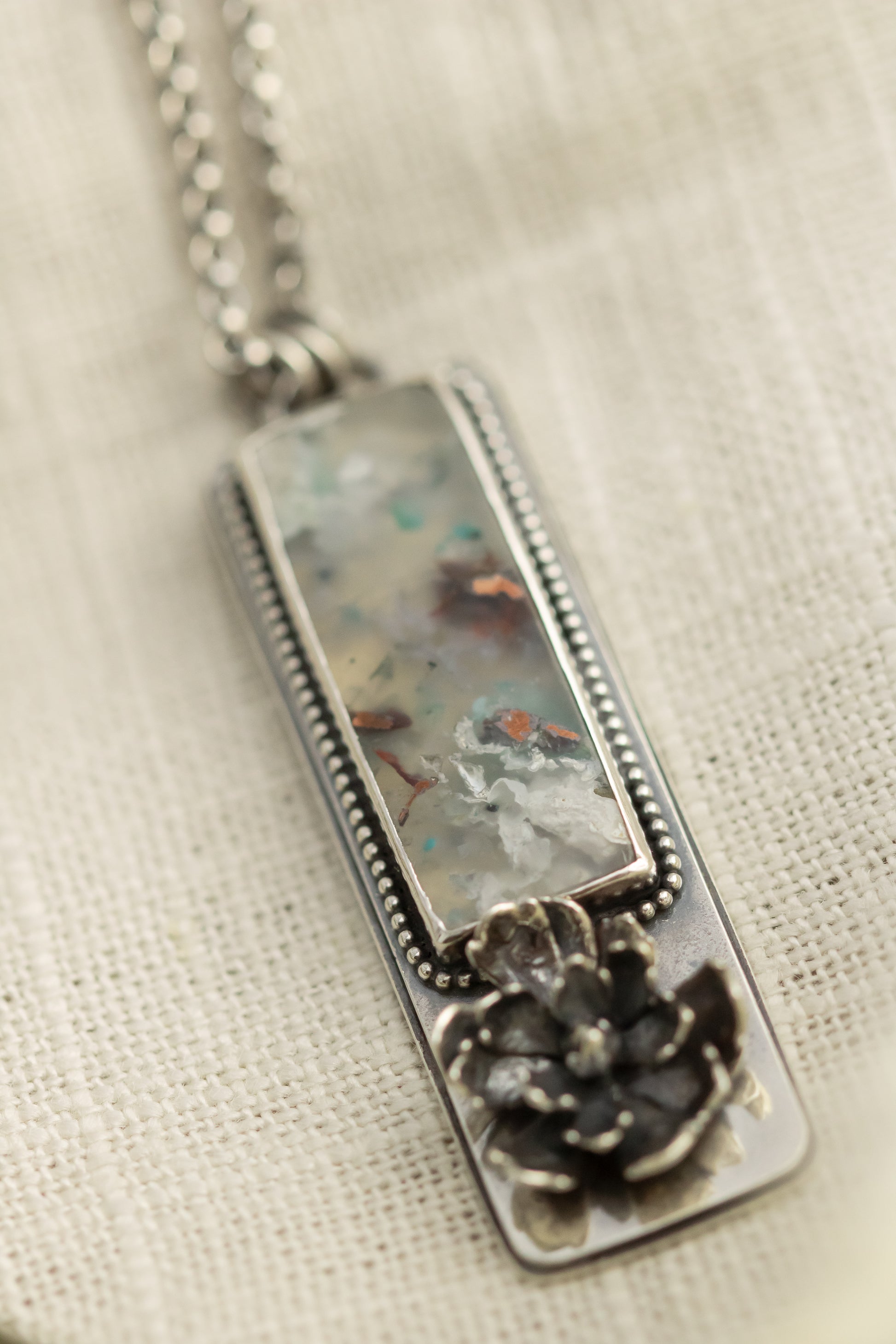 Sterling silver necklace with a sugar water flower agate pendant and succulent accent, laid flat on a beige fabric with dried eucalyptus leaves.
