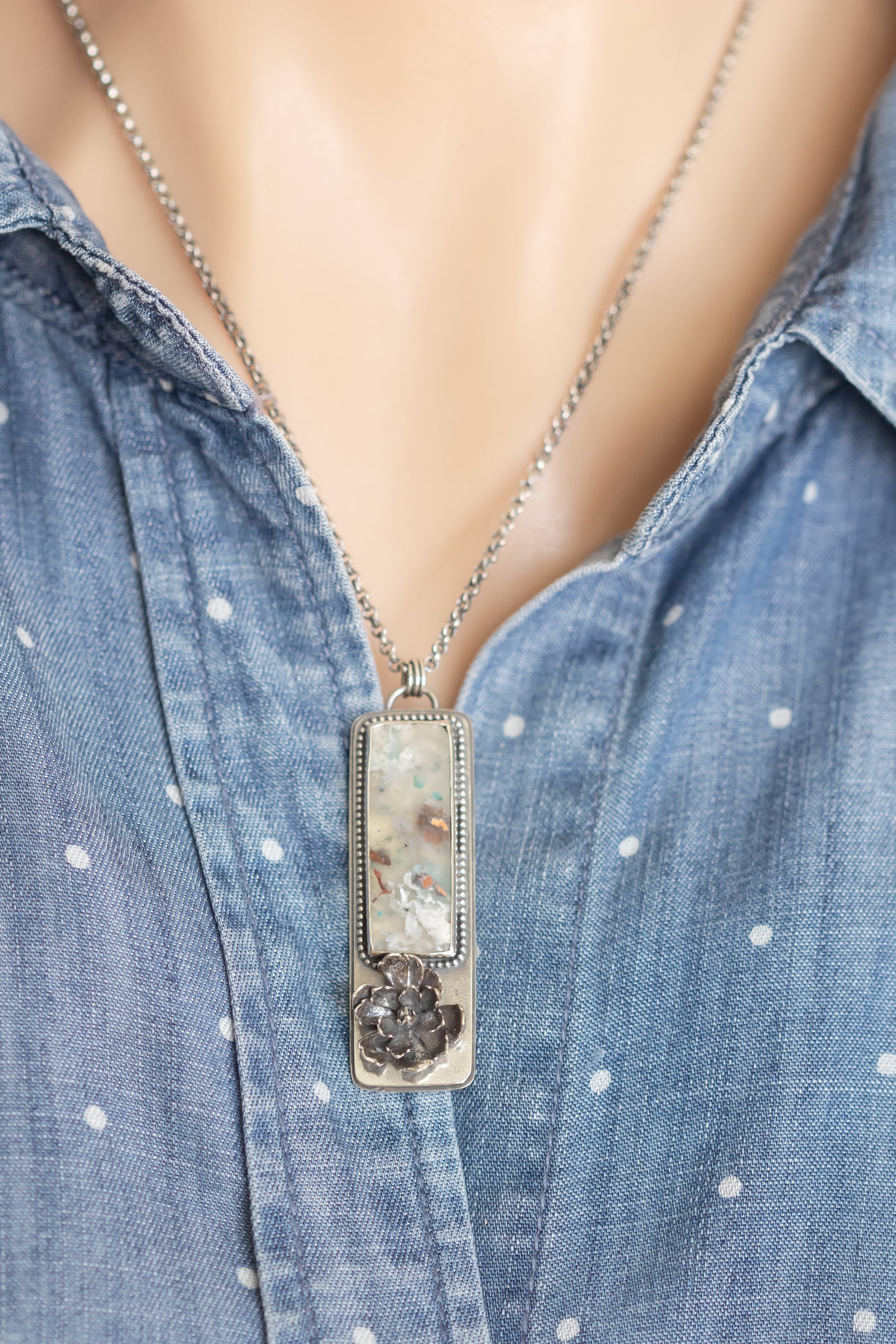 A sterling silver necklace with a sugar water flower agate pendant and succulent accent, worn on a model against a blue polka dot shirt.
