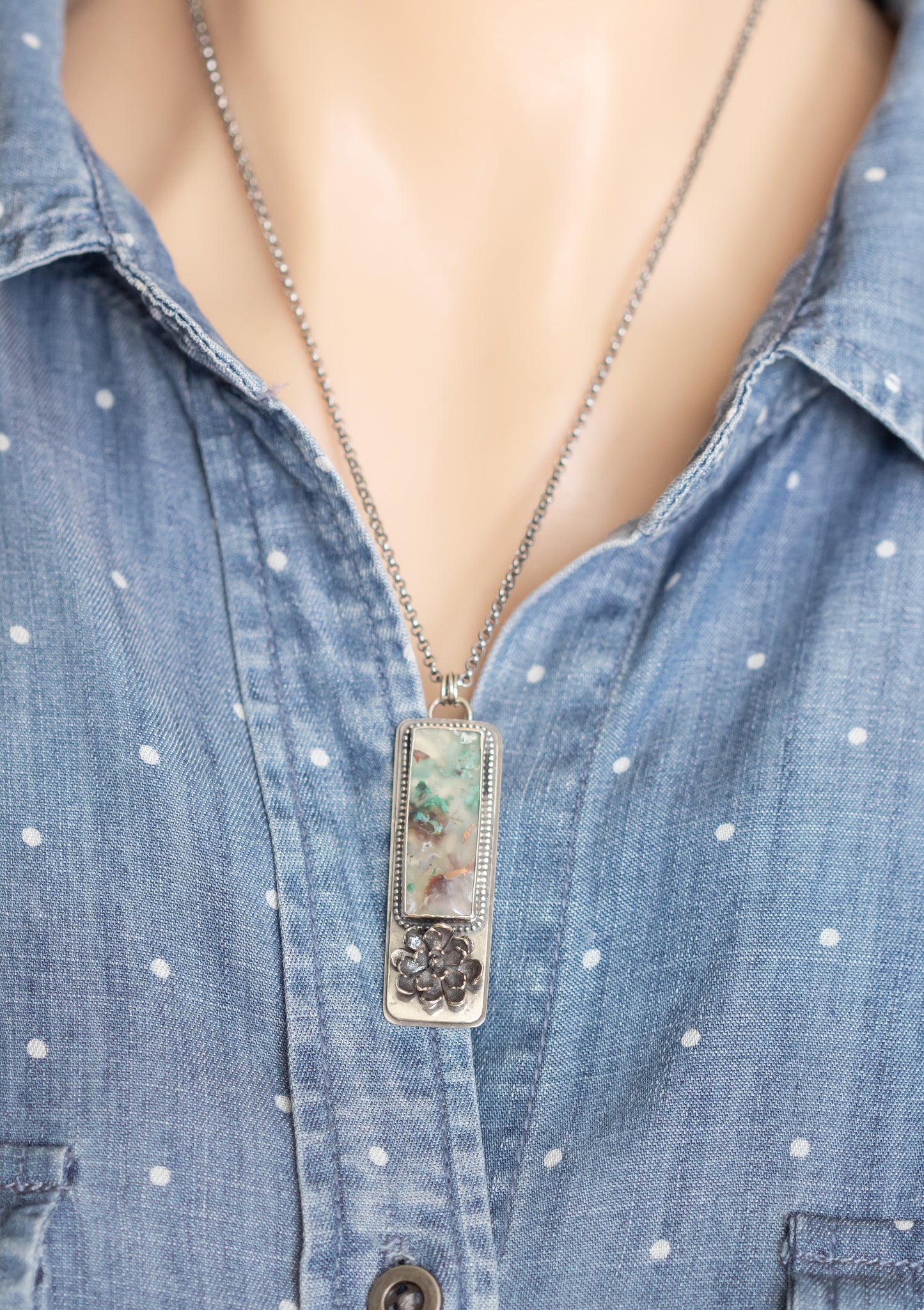  A sterling silver necklace with a sugar water flower agate pendant and succulent accent, worn on a model against a blue polka dot shirt.  
