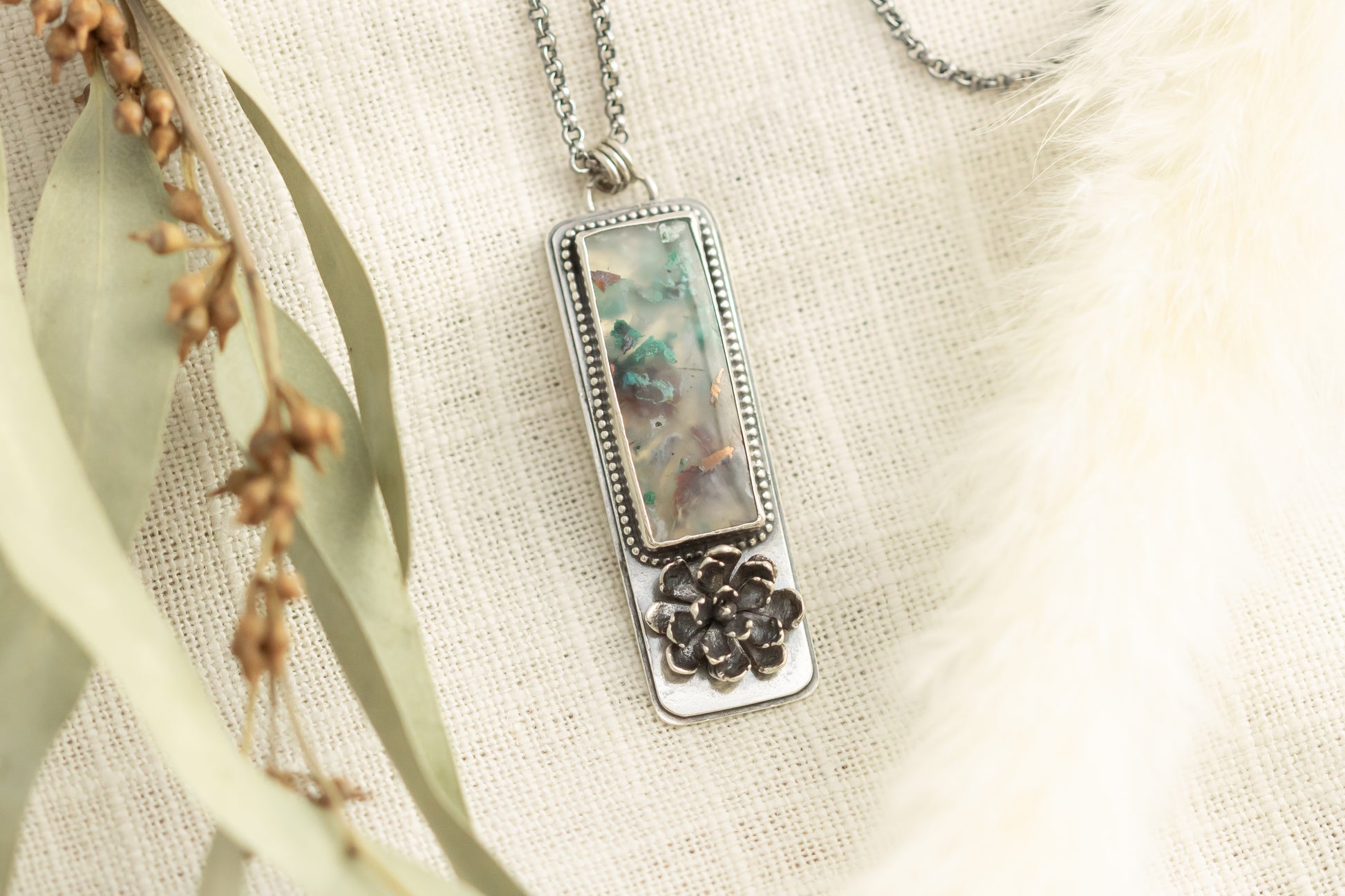 Sterling silver necklace with a sugar water flower agate pendant and succulent accent, laid flat on a beige fabric with dried eucalyptus leaves.
