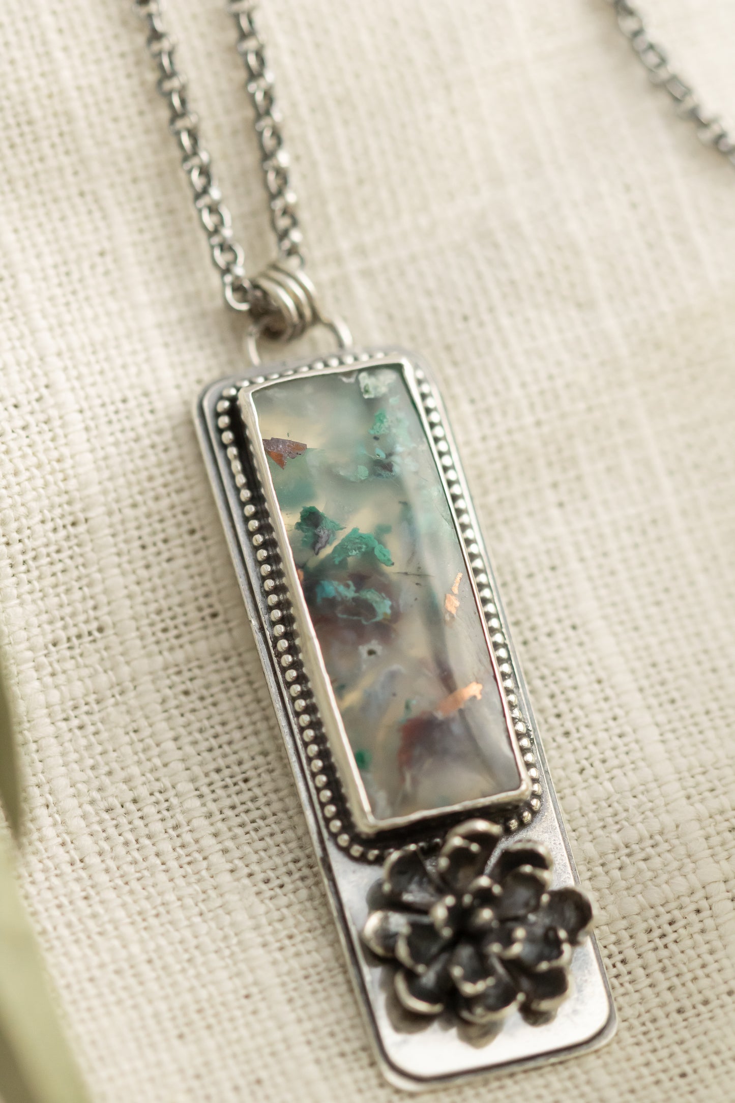Sterling silver necklace with a sugar water flower agate pendant and succulent accent, laid flat on a beige fabric with dried eucalyptus leaves.
