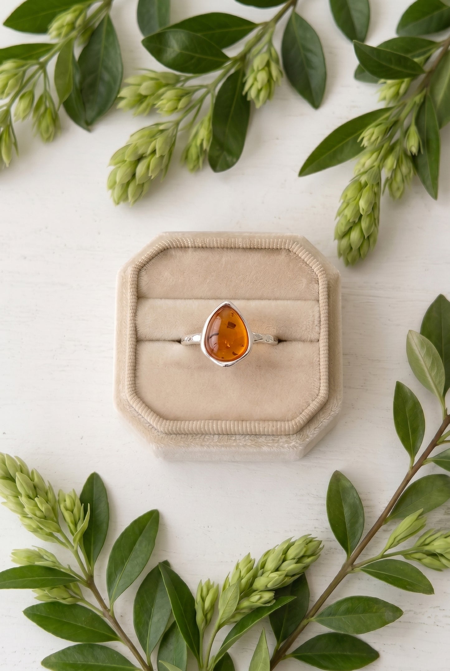 Flat lay view of a pear-shaped amber gemstone ring with a hammered sterling silver band sitting inside a light beige velvet jewelry box surrounded by green leafy branches.