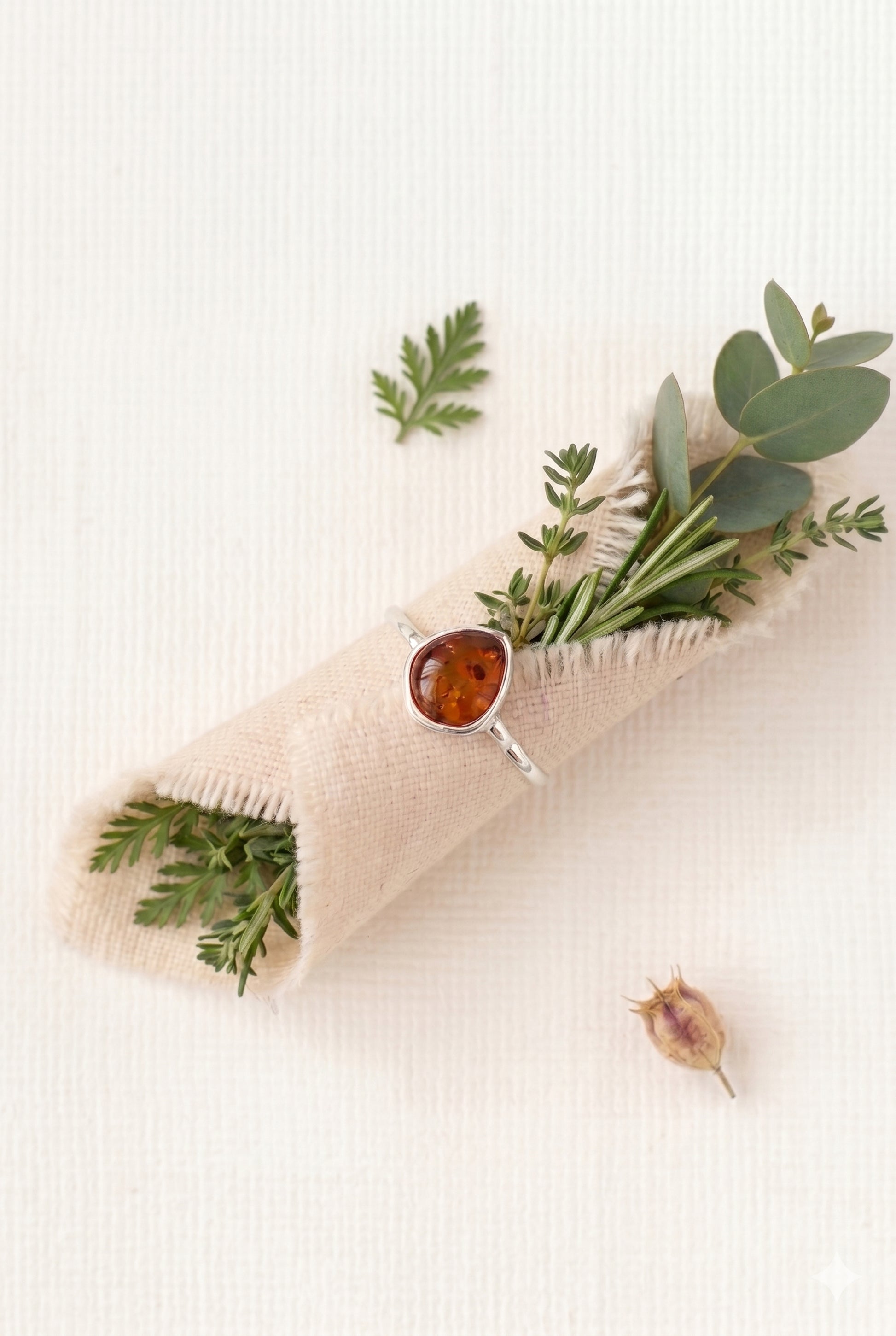 A teardrop-shaped amber stone ring with a sterling silver band wrapped around a rolled beige linen cloth containing sprigs of rosemary and eucalyptus on a white textured background.