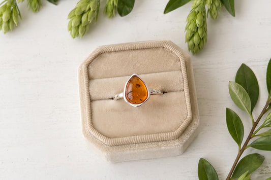 Flat lay view of a pear-shaped amber gemstone ring with a hammered sterling silver band sitting inside a light beige velvet jewelry box surrounded by green leafy branches.