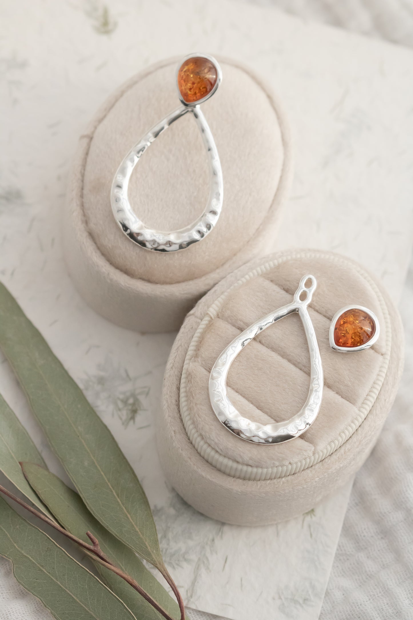 Sterling silver hammered pear drop Baltic amber dangle convertible stud earrings displayed on two beige velvet jewelry pedestals with fresh eucalyptus leaves  
