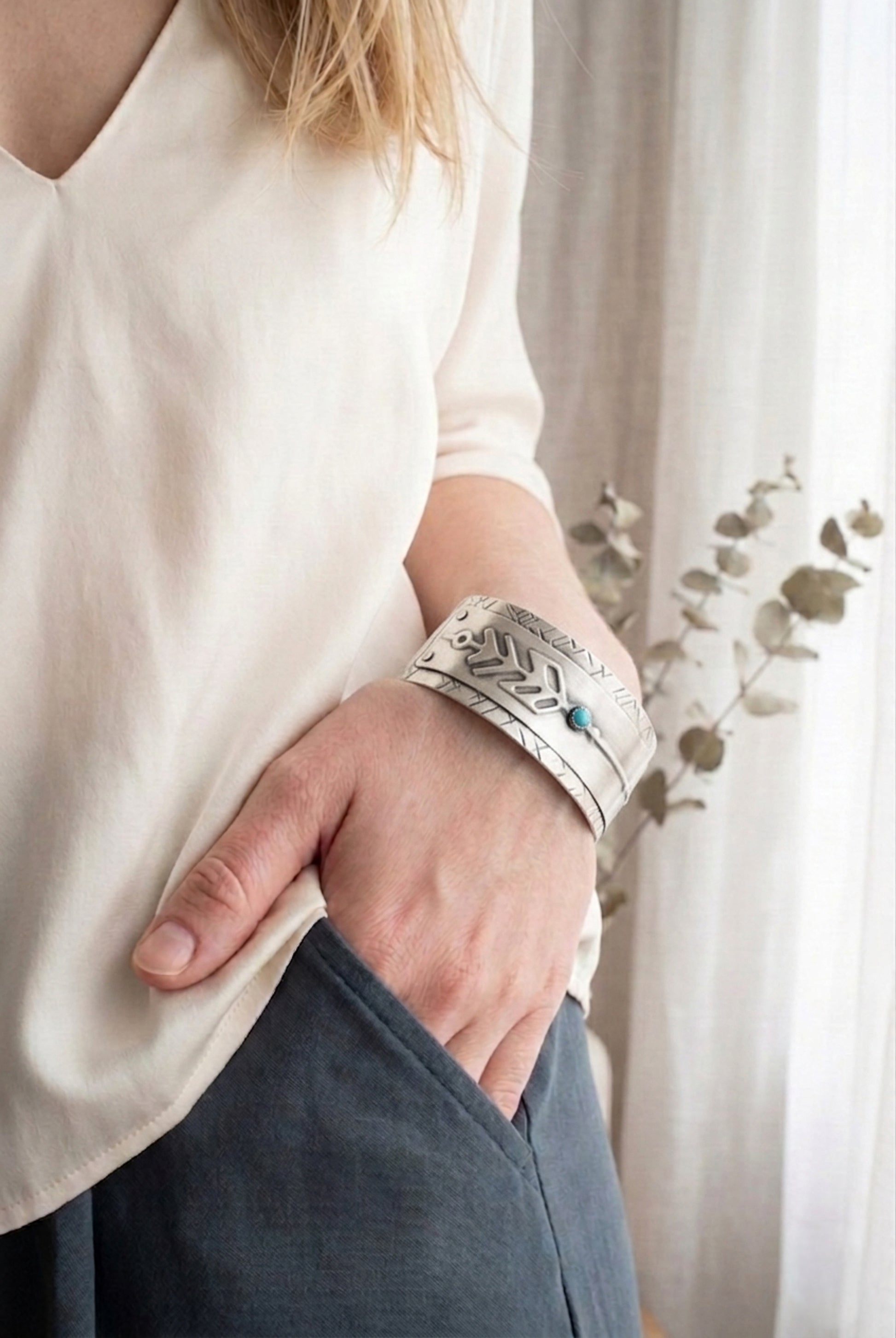Person wearing a sterling silver cuff bracelet with an arrow design and turquoise stone wearing a white shirt and dark pants, standing against a neutral background.