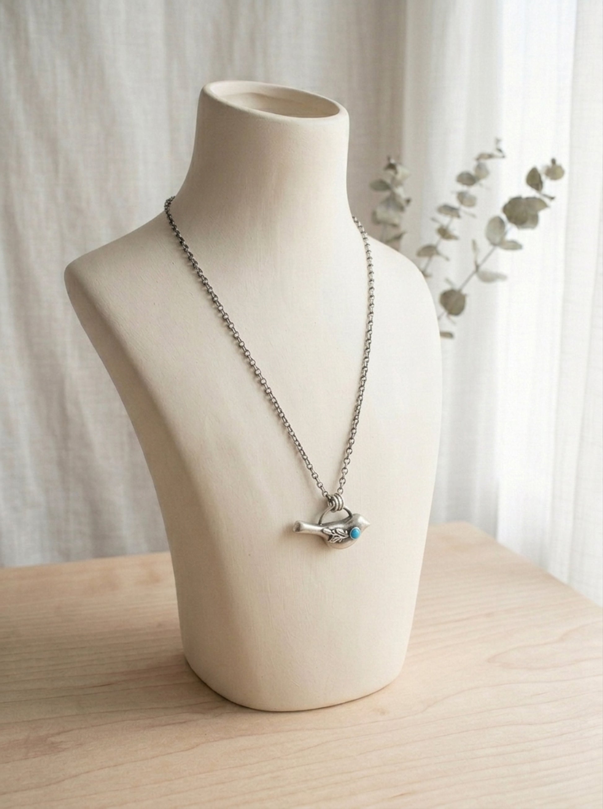 A sterling silver necklace with a bird pendant featuring a turquoise gemstone and a delicate leaf design on a display bust. 