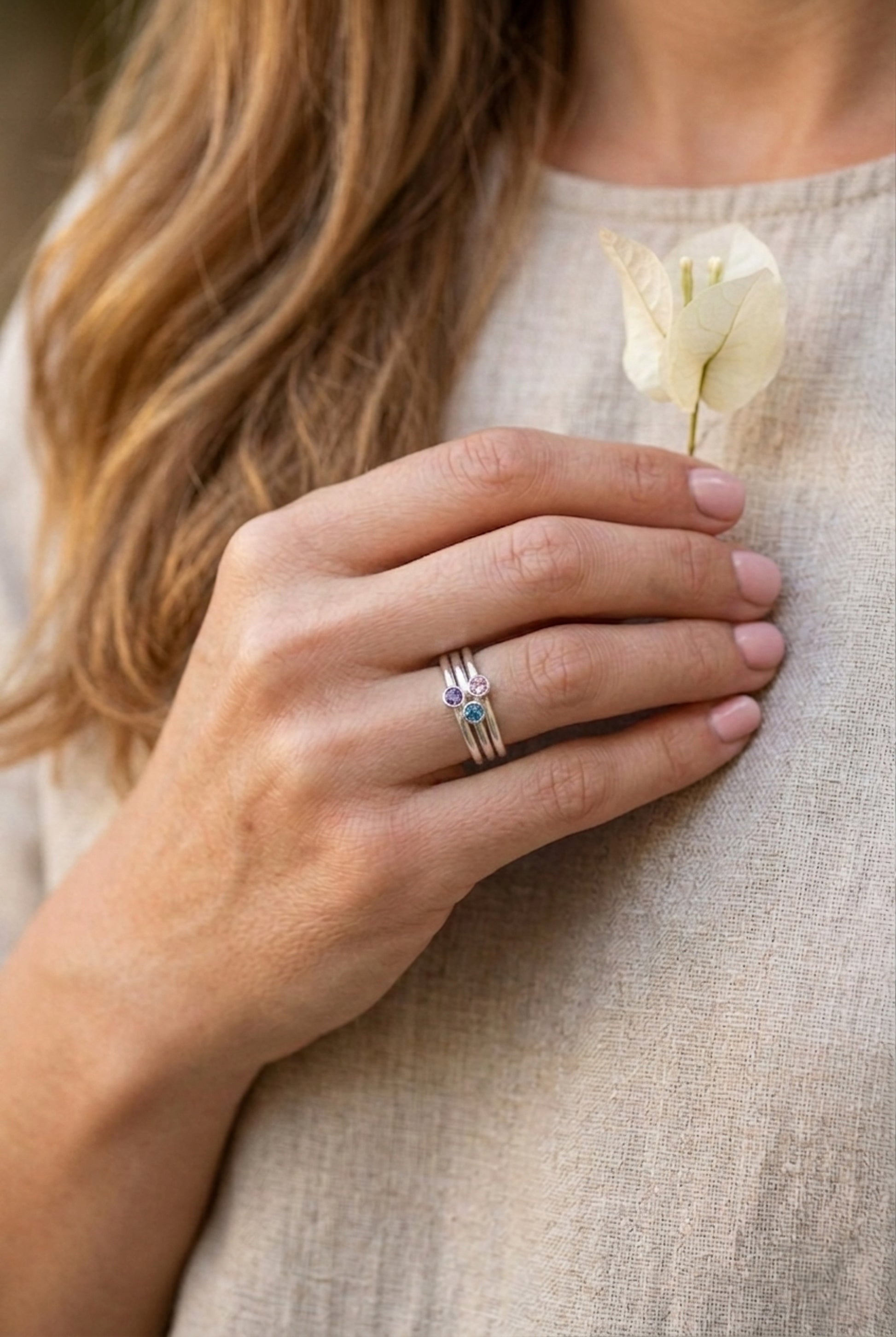 Lifestyle shot of a woman with long, light-brown wavy hair. She is wearing a beige, linen-textured top. Her right hand, with light pink fingernails, is held gently against her chest. She is wearing the stacked three-stone birthstone ring set from (with teal, pink, and purple stones) on her ring finger. Her index finger and thumb are lightly holding a single, delicate white bougainvillea blossom. The background is softly blurred.