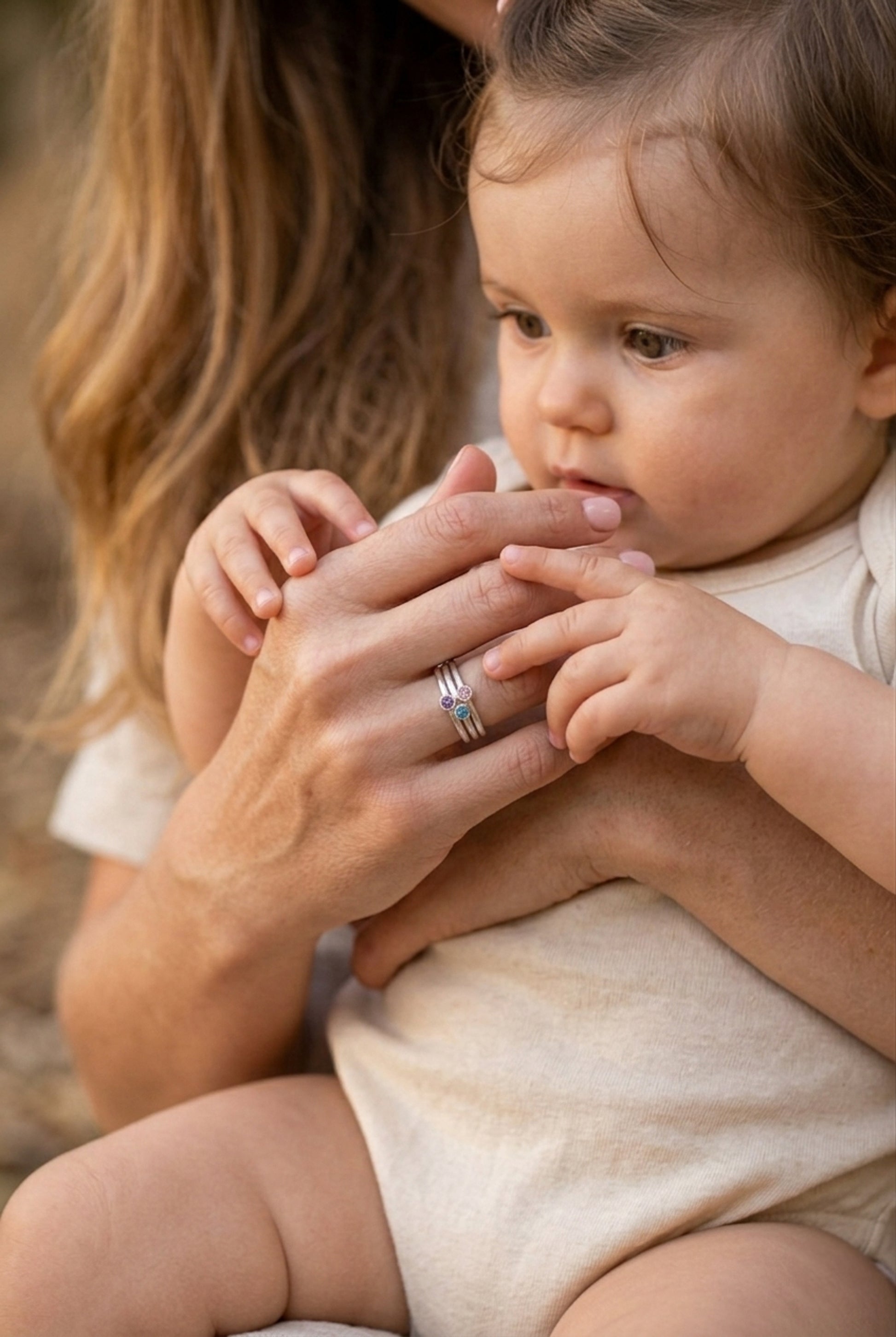 heartwarming close-up of an adult and baby's hands. The adult's right hand, with a polished light pink manicure, is in the foreground, wearing the stacked three-stone birthstone ring set (teal, pink, purple gems). Her hand is being tenderly held and explored by the soft, chubby hand of a baby. A partial view of the adult's blonde hair is on the left, and the baby's cheek is softly in focus on the right. The lighting is warm and natural.