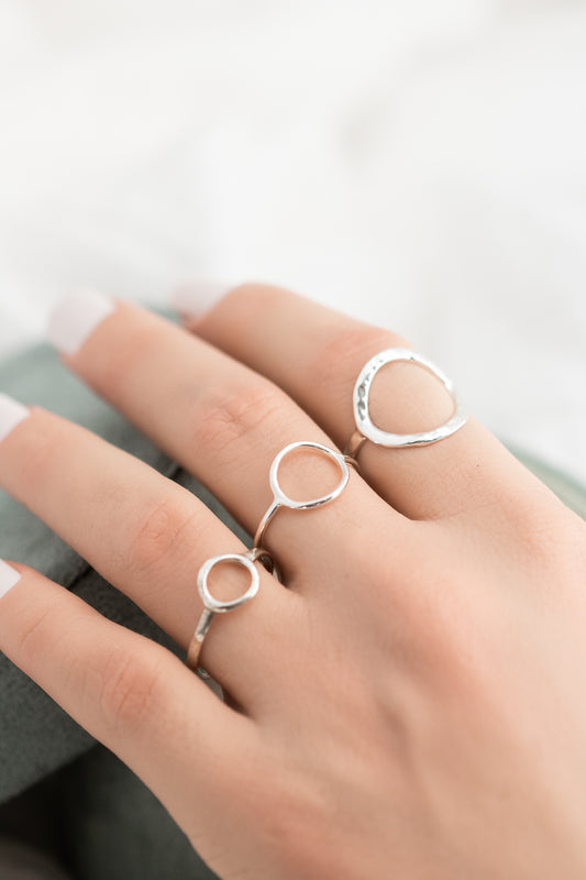 A close-up of a hand wearing three open circle sterling silver rings in small, medium, and large sizes, with the large ring featuring a hammered finish, against a soft background.
