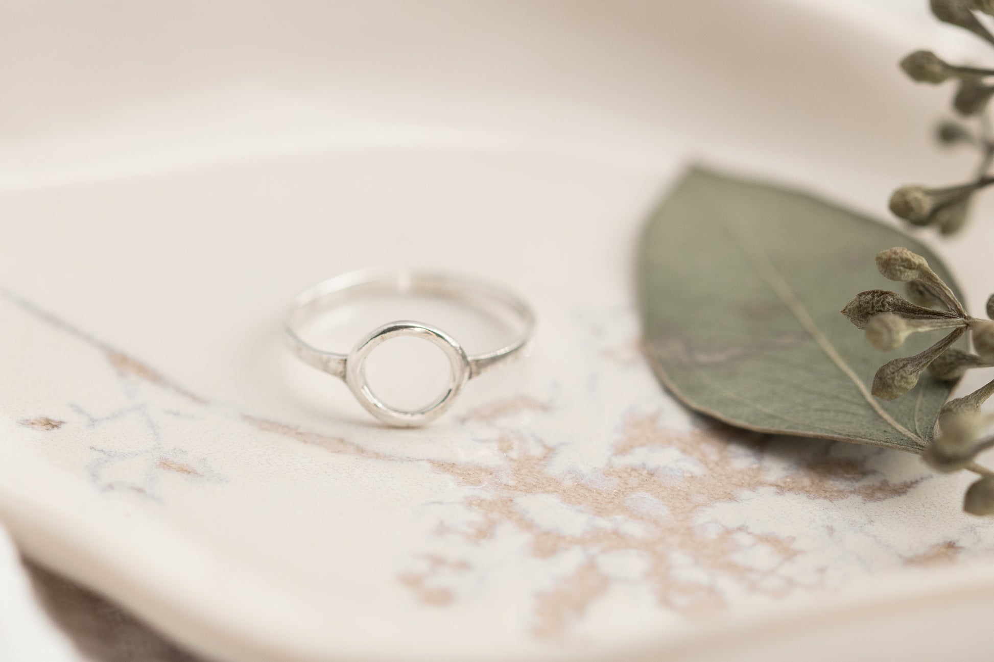  A single open circle sterling silver ring on a white marbled dish with a green leaf, set against a soft white fabric background.

