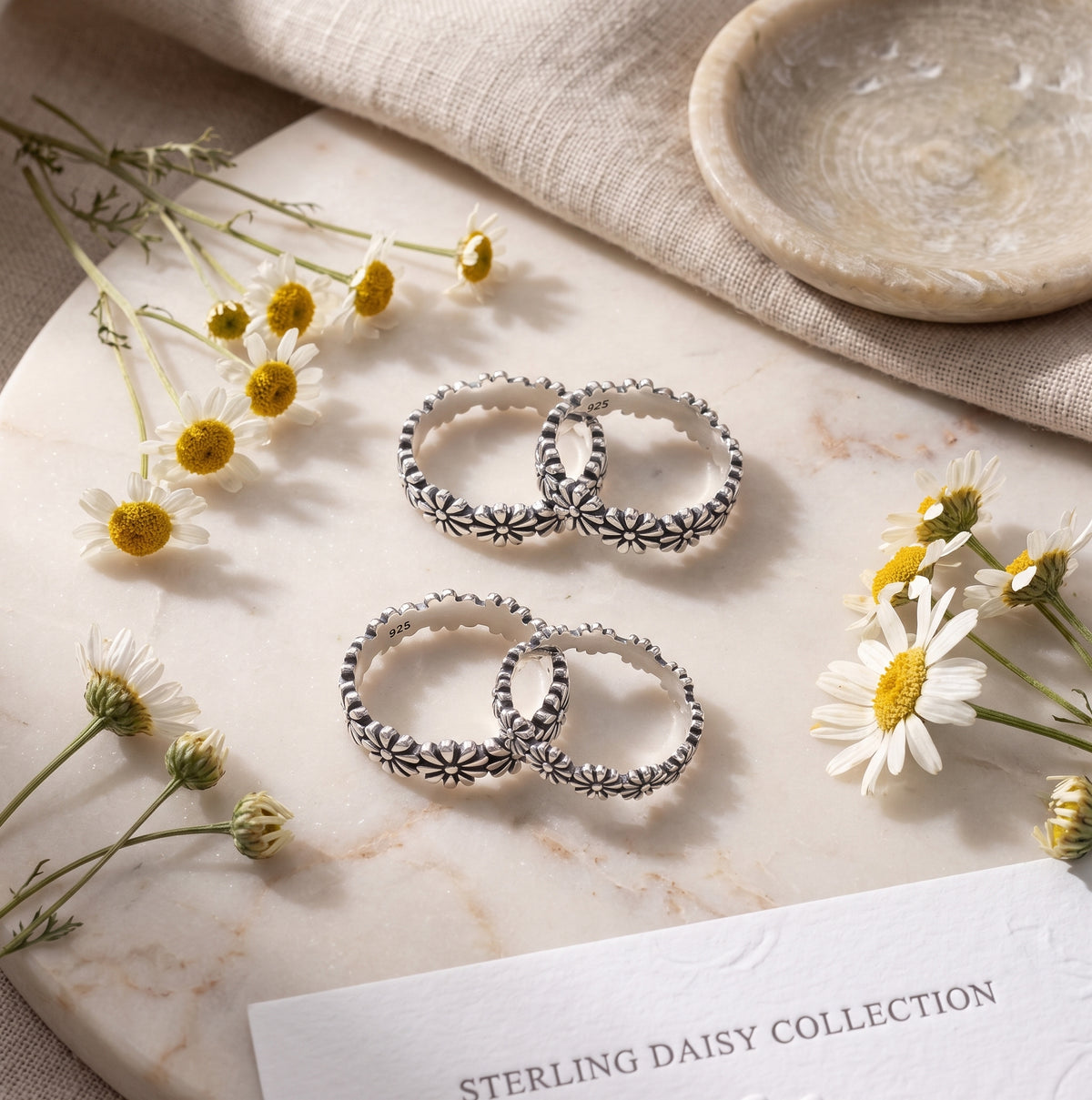 Stack of sterling silver daisy floral bands on a marble surface surrounded by fresh white daisies and soft linen backdrop.