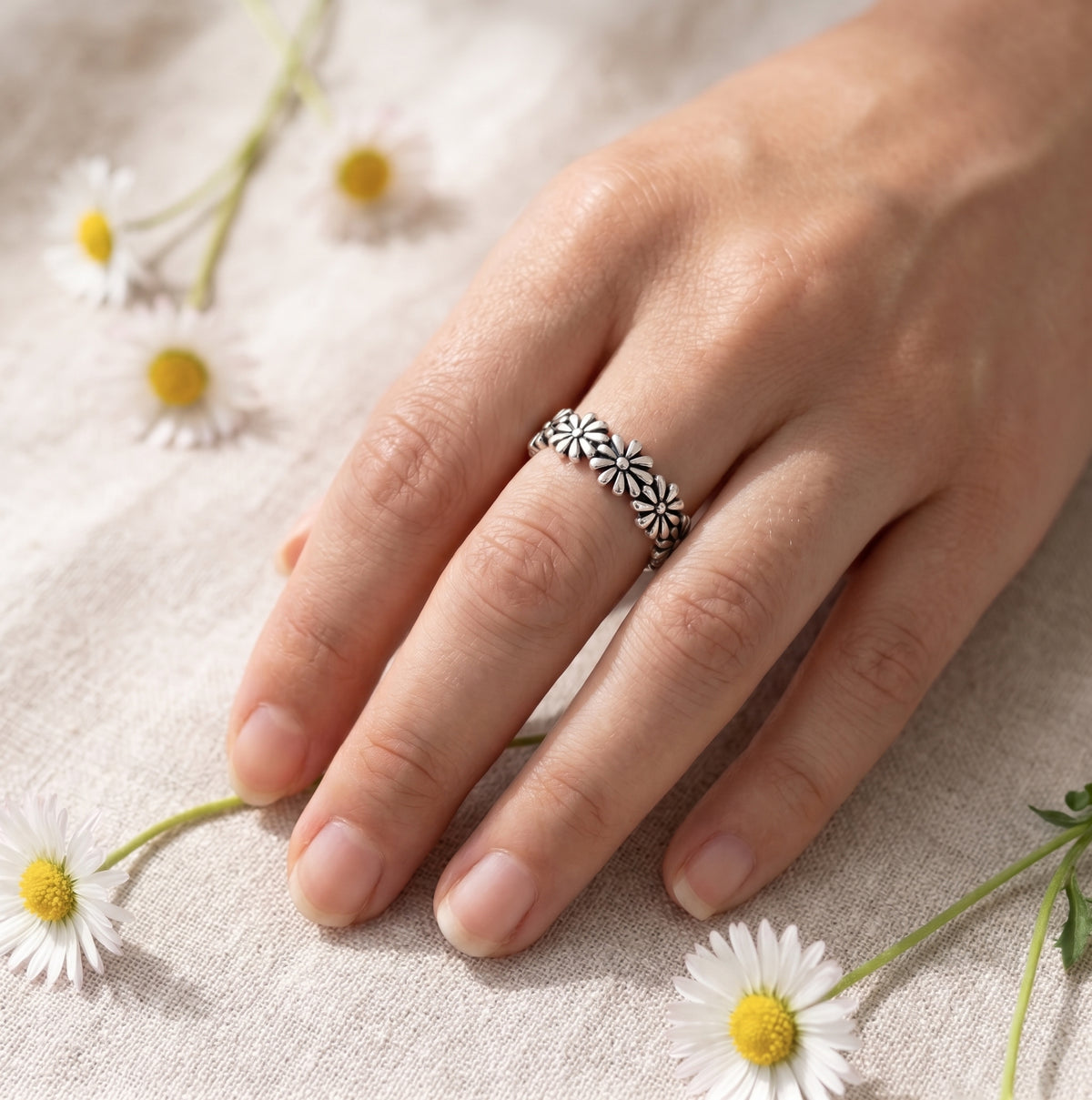Hand modeling a dainty oxidized sterling silver daisy flower band worn on middle finger over beige linen texture.