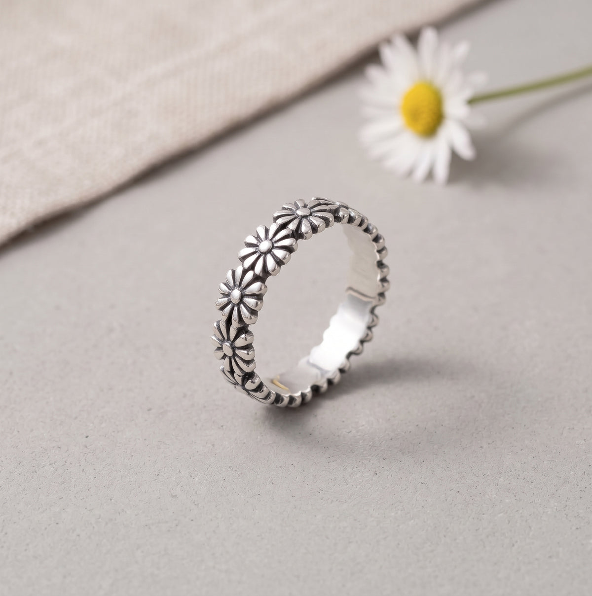 Sterling silver daisy band ring featuring detailed floral engravings on a neutral stone background with fresh daisy accents.