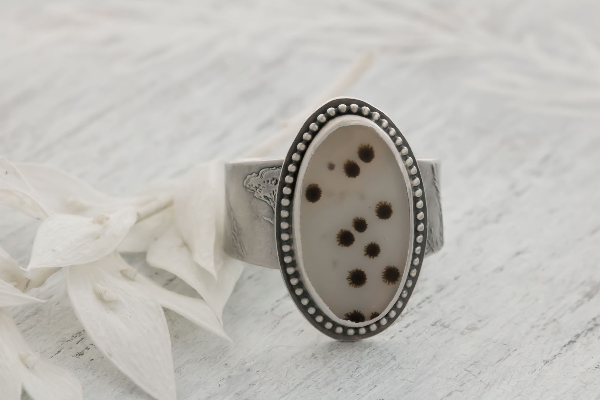 A handmade sterling silver dendritic agate ring resting on a light textured surface next to a white petal. The large oval cabochon displays scattered black and brown dendritic inclusions like tiny polka dots or fireworks on a whitish-gray stone, accented by a dotted bezel and engraved wildflower/wheatgrass band. Perfect bohemian nature jewelry.
