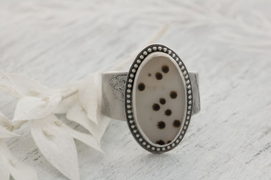 A handmade sterling silver dendritic agate ring resting on a light textured surface next to a white petal. The large oval cabochon displays scattered black and brown dendritic inclusions like tiny polka dots or fireworks on a whitish-gray stone, accented by a dotted bezel and engraved wildflower/wheatgrass band. Perfect bohemian nature jewelry.

