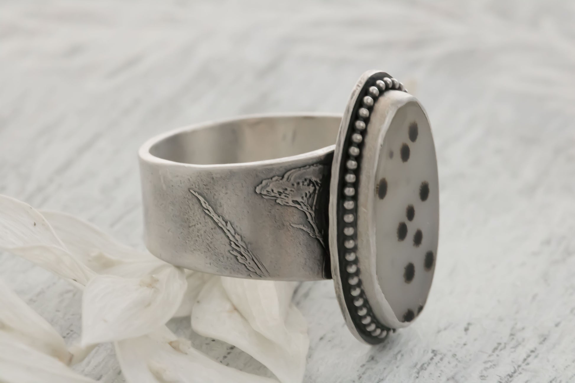 A handmade sterling silver dendritic agate ring resting on a light textured surface next to a white petal. The large oval cabochon displays scattered black and brown dendritic inclusions like tiny polka dots or fireworks on a whitish-gray stone, accented by a dotted bezel and engraved wildflower/wheatgrass band. Perfect bohemian nature jewelry.
