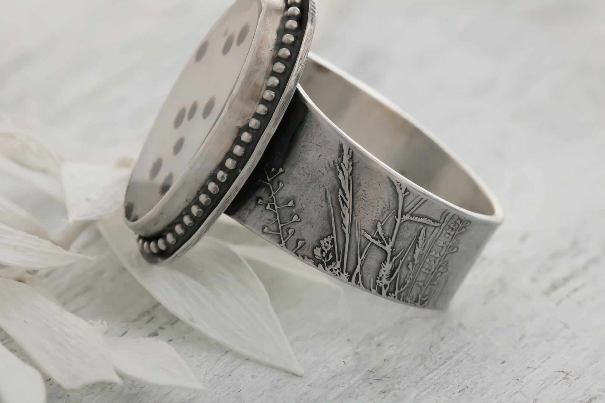 A handmade sterling silver dendritic agate ring resting on a light textured surface next to a white petal. The large oval cabochon displays scattered black and brown dendritic inclusions like tiny polka dots or fireworks on a whitish-gray stone, accented by a dotted bezel and engraved wildflower/wheatgrass band. Perfect bohemian nature jewelry.

