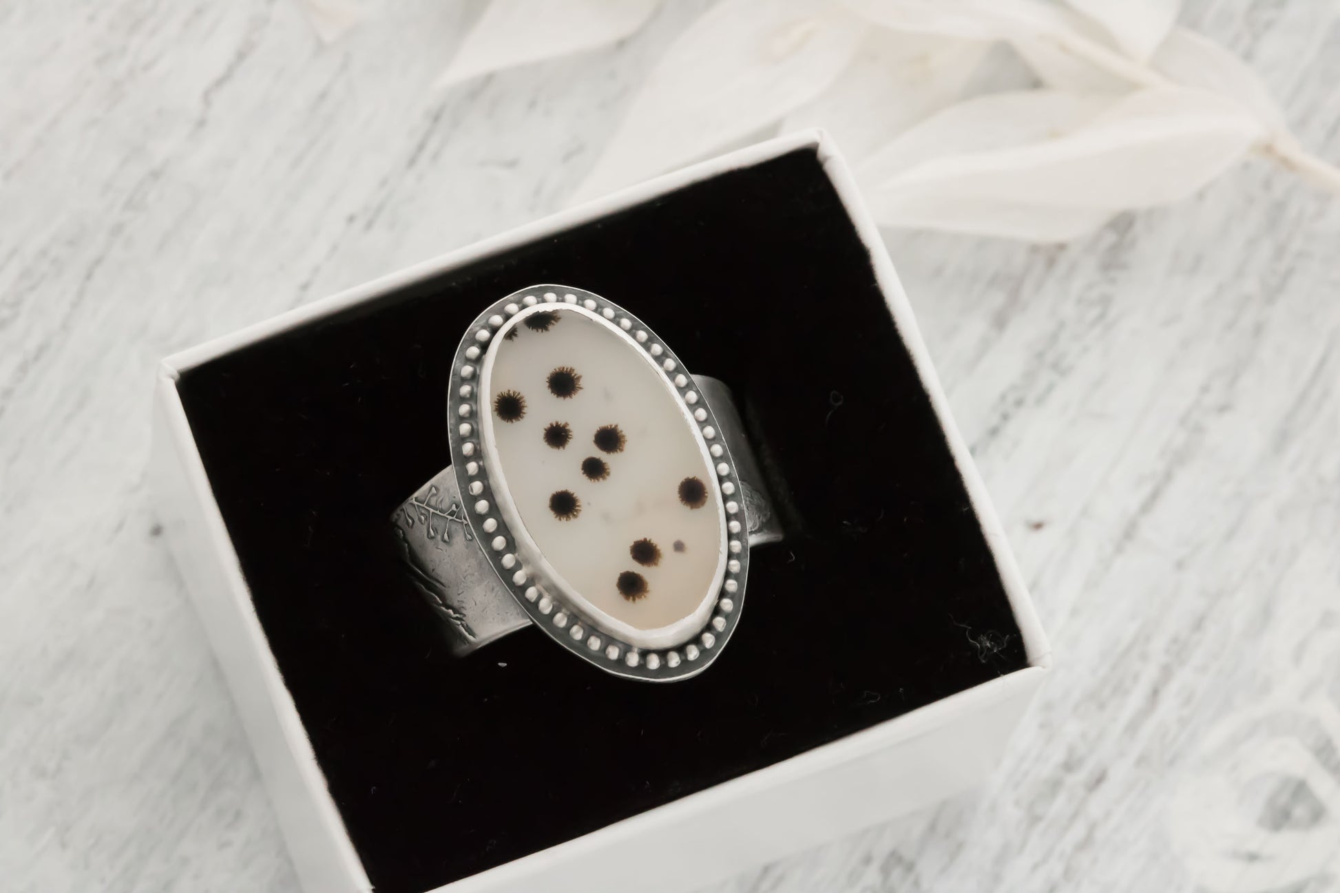  Handcrafted sterling silver dendritic agate ring displayed in an open black jewelry box on a soft white marble background. The oval cabochon stone features unique black and brown fern-like dendritic inclusions resembling nature's polka dots on a translucent whitish-gray base, with a beaded bezel and wildflower/wheatgrass textured band. Size 9, one-of-a-kind artisan piece from Alaska.
