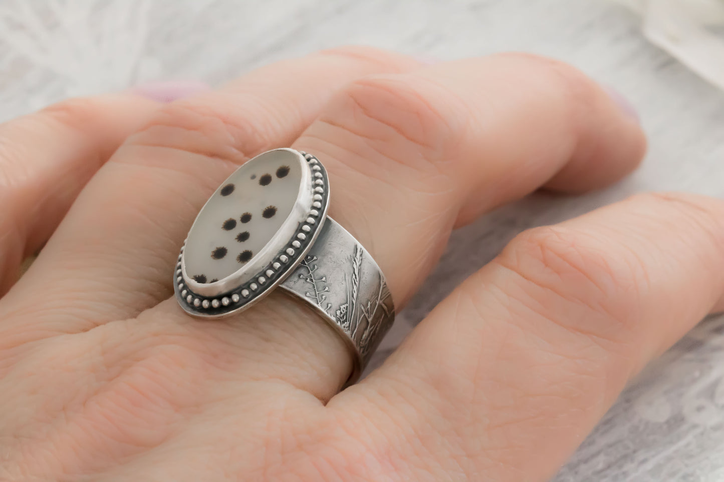 Close-up of a hand wearing a size 9 handcrafted dendritic agate ring in sterling silver. The oval stone shows distinctive black branch-like dendrite patterns on a milky translucent background, framed by a beaded bezel setting. The wide band is textured with delicate wildflower and wheatgrass impressions for a rustic, nature-inspired look.

