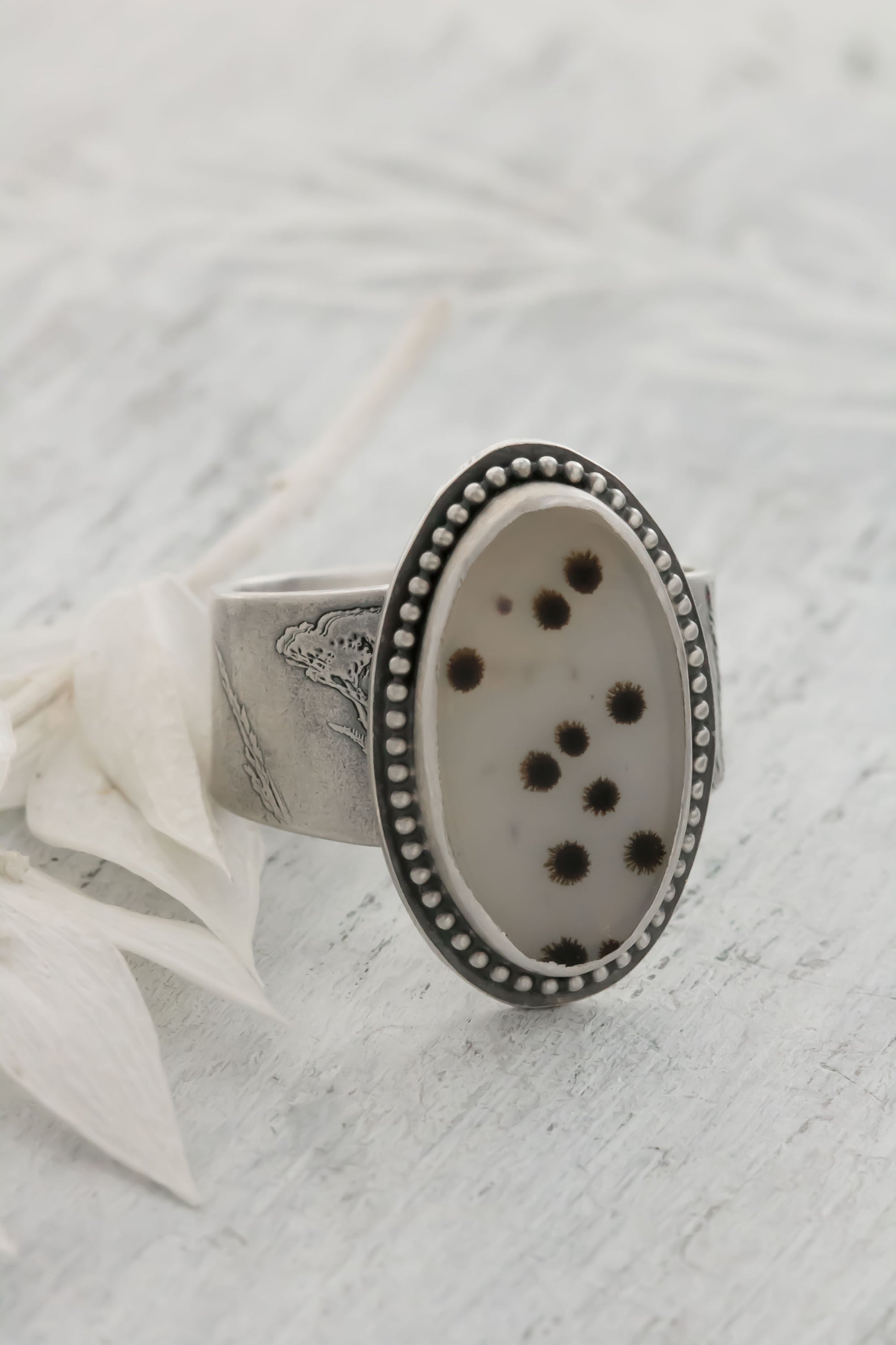 A handmade sterling silver dendritic agate ring resting on a light textured surface next to a white petal. The large oval cabochon displays scattered black and brown dendritic inclusions like tiny polka dots or fireworks on a whitish-gray stone, accented by a dotted bezel and engraved wildflower/wheatgrass band. Perfect bohemian nature jewelry.
