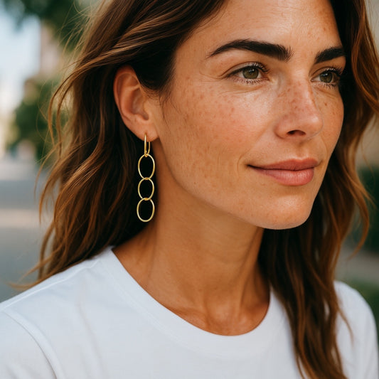 A woman with wavy brown hair wearing 18K yellow gold hammered triple hoop earrings, outdoors with a blurred green background, in a white top.  
