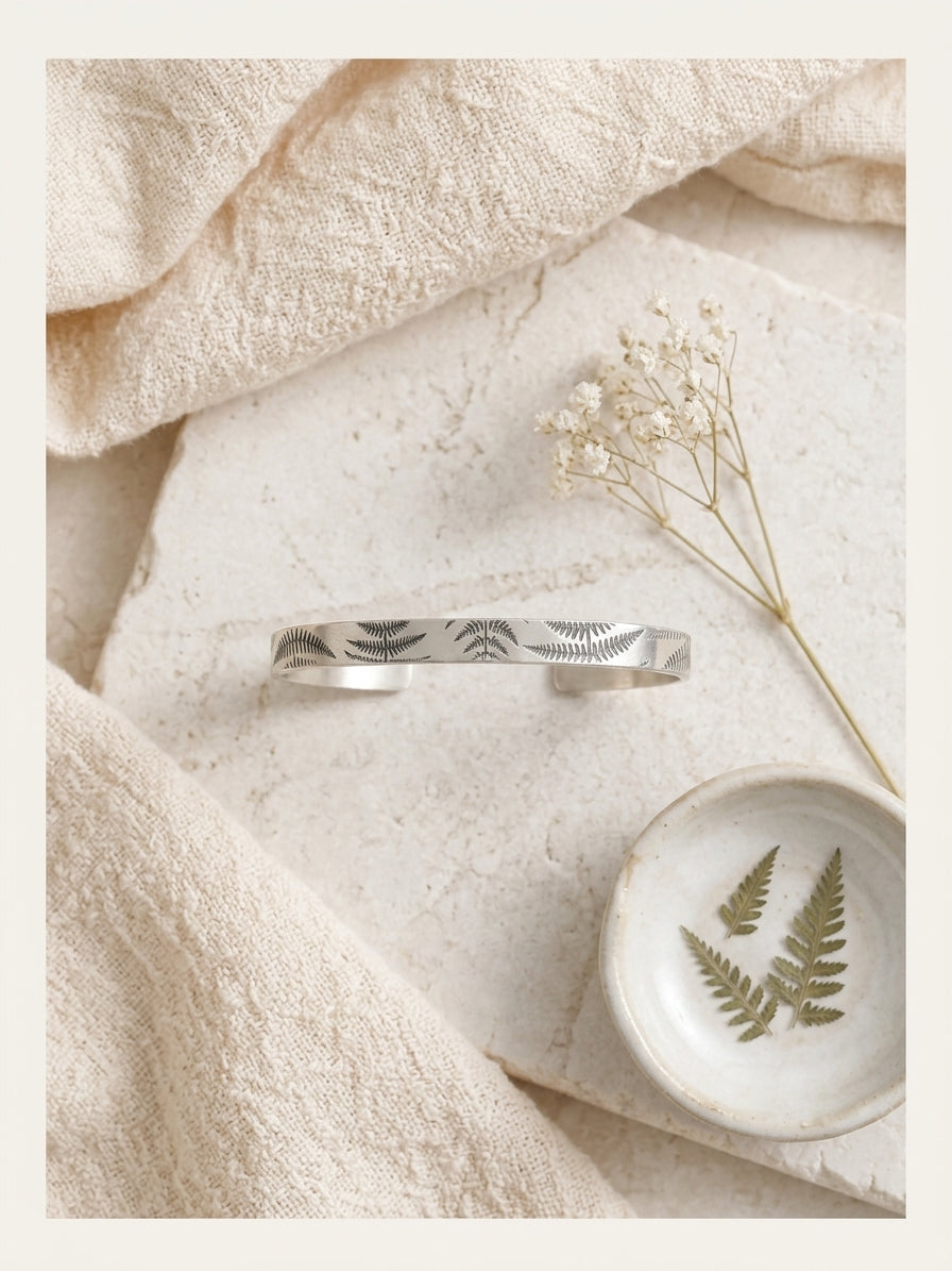 A sterling silver cuff bracelet with engraved fern leaf patterns, styled in a flat-lay arrangement on a textured stone slab with beige linen, dried baby's breath, and a small dish with pressed fern leaves.