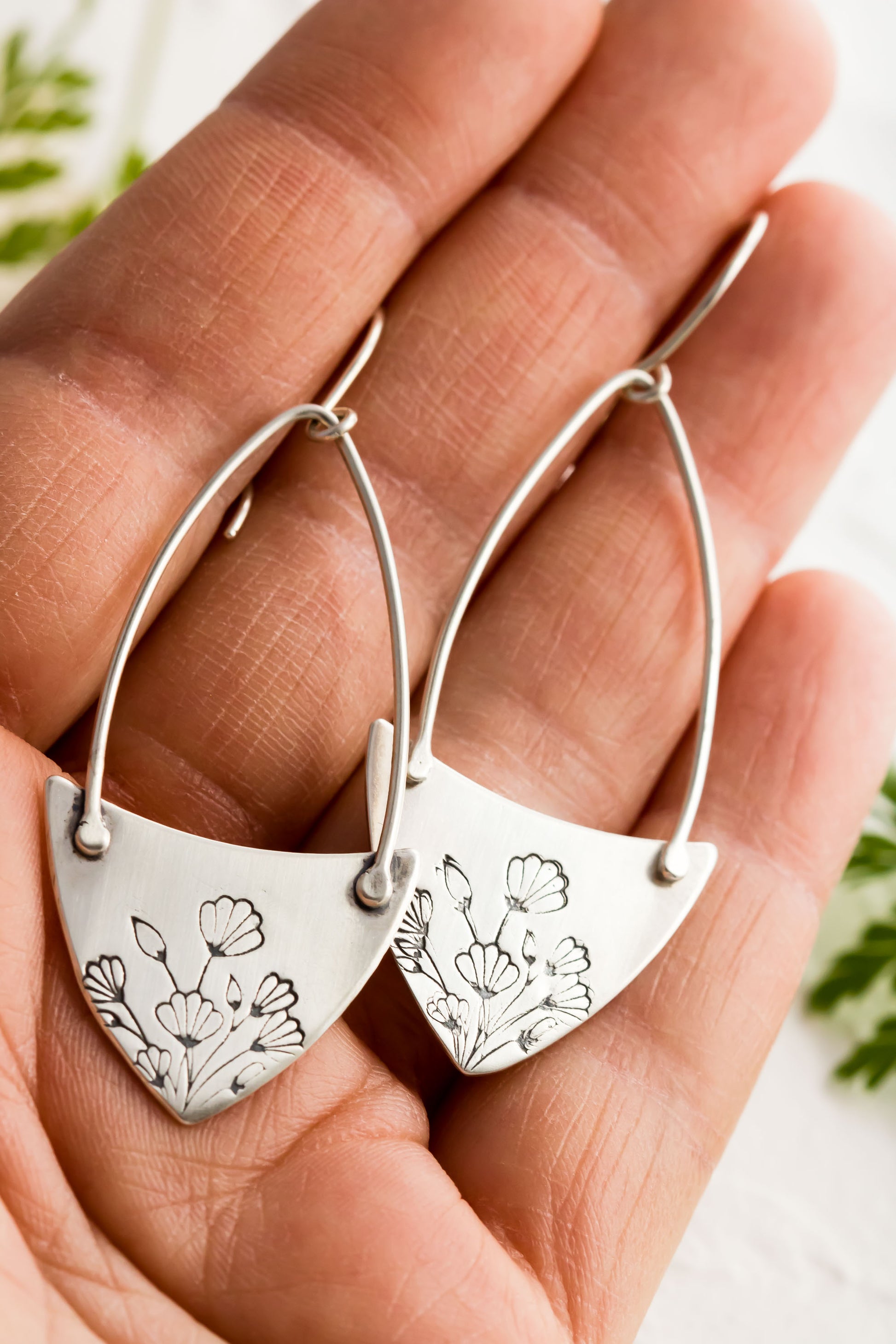 Handcrafted sterling silver Art Nouveau floral hoop earrings held I a hand with a textured background with greenery. 
