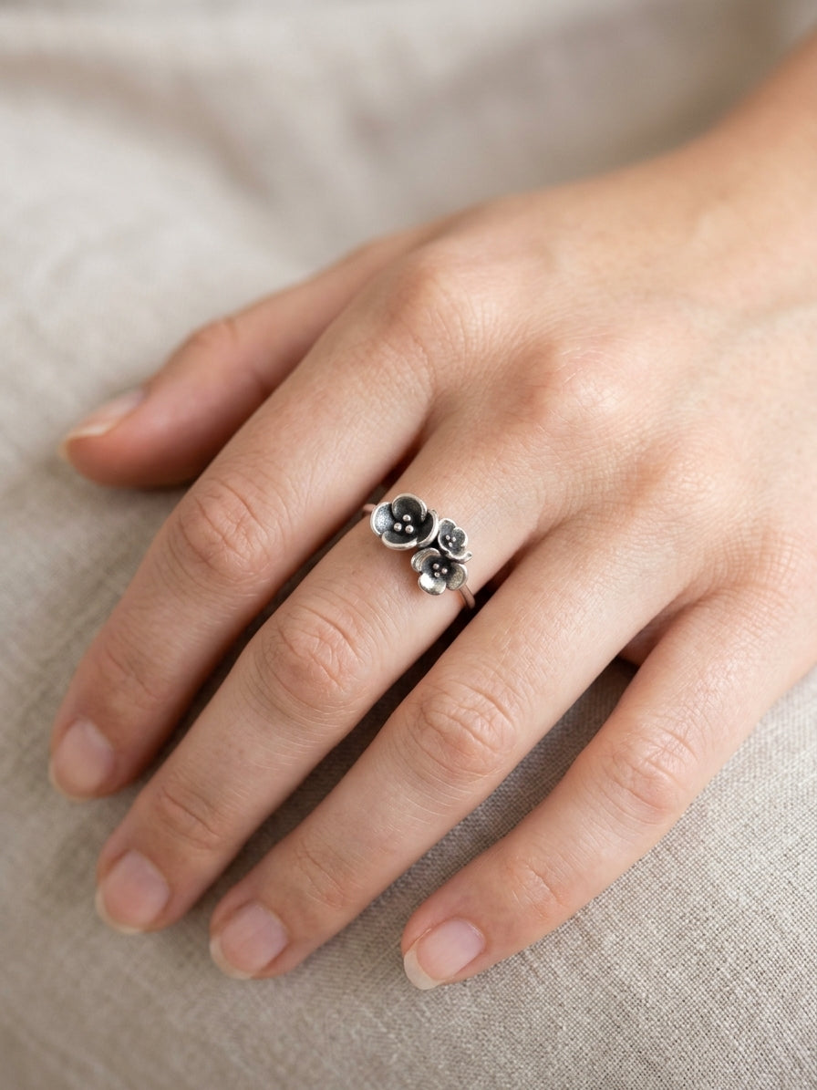 A close-up of a hand wearing a  sterling silver ring with three small  flowers on a soft beige fabric background.