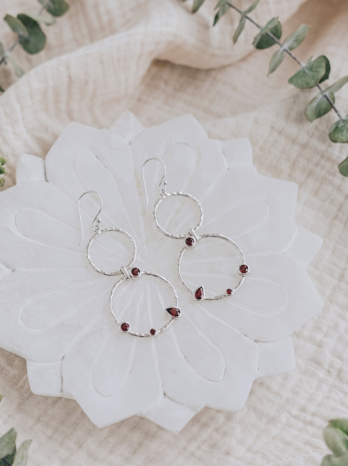 Hammered silver double-circle earrings with round and pear-shaped red garnets on a white flower-shaped dish.