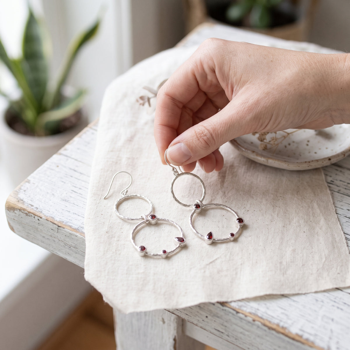 Hand holding one garnet double-circle drop earring above cream fabric, with the matching earring resting beside it.
