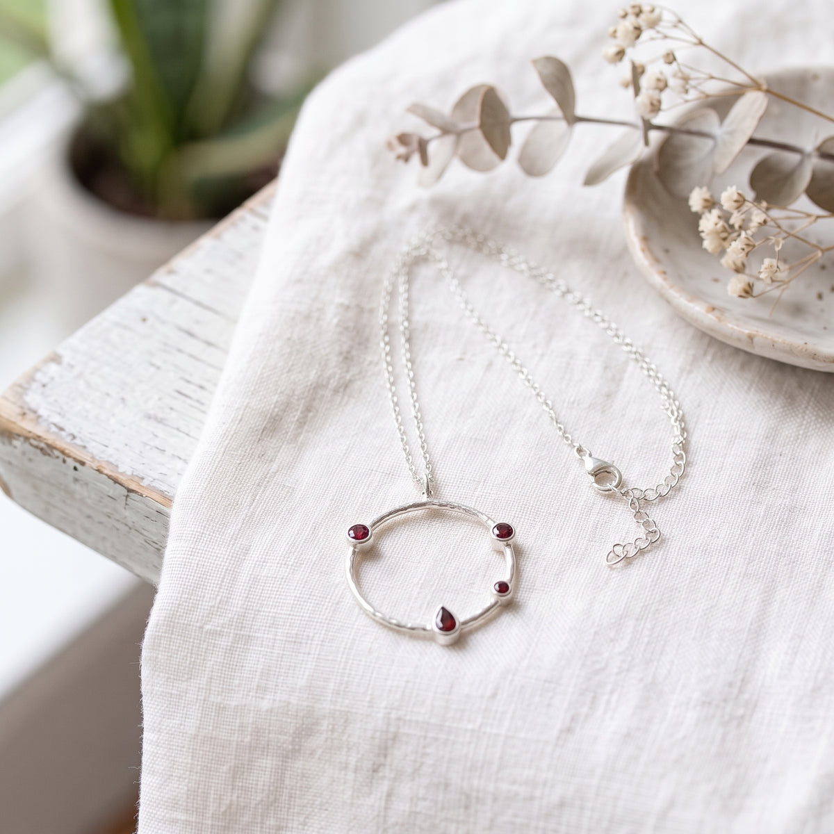 Garnet hammered circle necklace on white fabric, showing round and pear-shaped stones, silver chain, lobster clasp and extender.