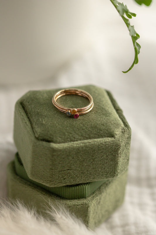 14K yellow gold birthstone stackable rings with 2mm round stones on a green velvet box, white fabric, and green plant in the background.
