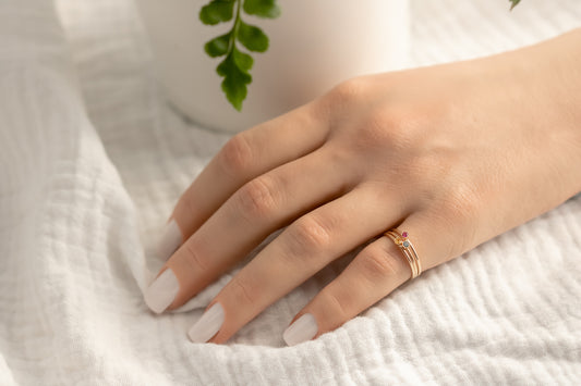 Hand wearing 14K yellow gold birthstone stackable rings with 2mm round stones, sizes 4-10, on white fabric with green plant in background.
