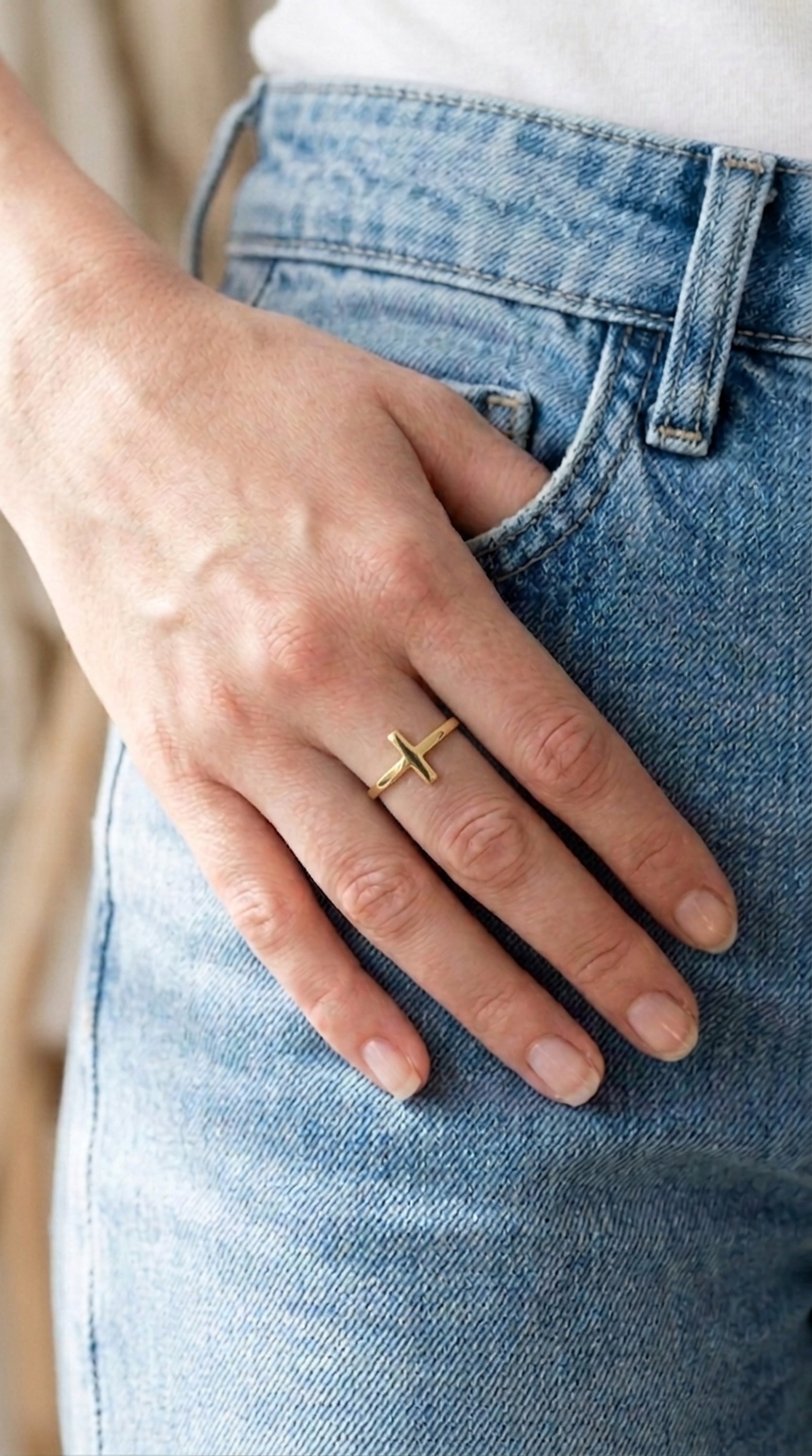 A close-up of a hand resting on a denim pocket wearing a minimalist 14k gold horizontal cross ring. The sideways cross ring features a thin, polished band and a sleek religious symbol, perfect for casual everyday wear or a modern christian jewelry style.