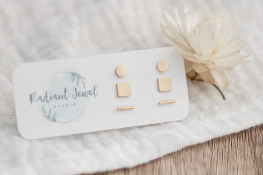 Close-up of 14k gold filled stud earring set on branded card with flower, showing circle, square, and bar shapes.
