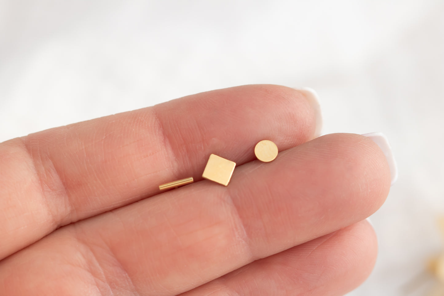 Hand holding tiny 14k gold filled stud earrings in bar, square, and circle designs against white background.
