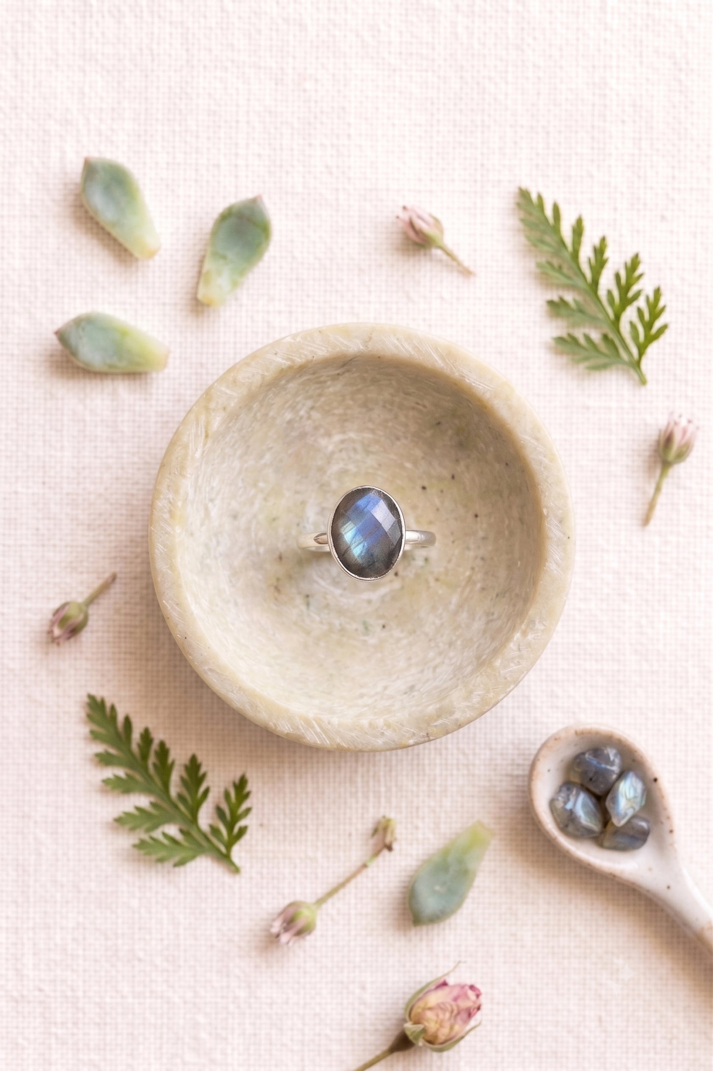 Statement labradorite ring with a faceted oval stone resting inside a small carved beige stone bowl. Boho jewelry flat lay with scattered botanical leaves, flower buds, and raw crystals.