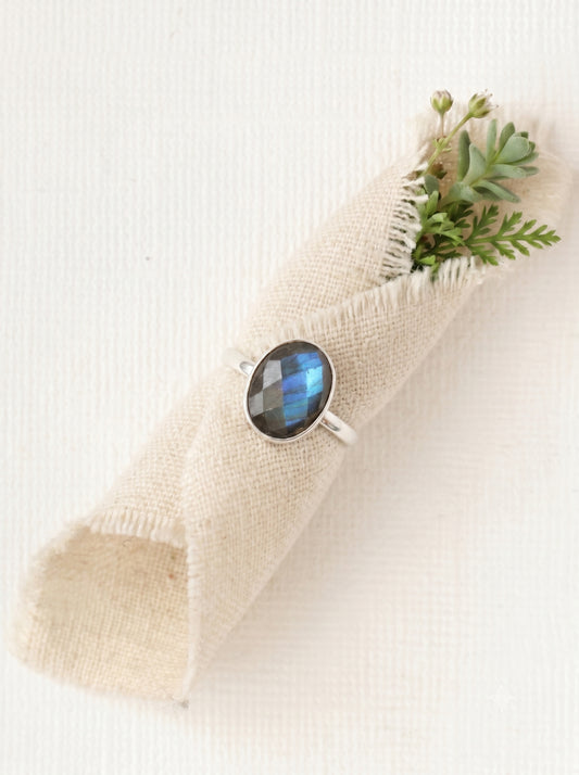 Sterling silver labradorite gemstone ring wrapped around a rustic cream linen fabric roll with green succulents. High-quality oval statement ring showcasing a vibrant blue fire flash.
