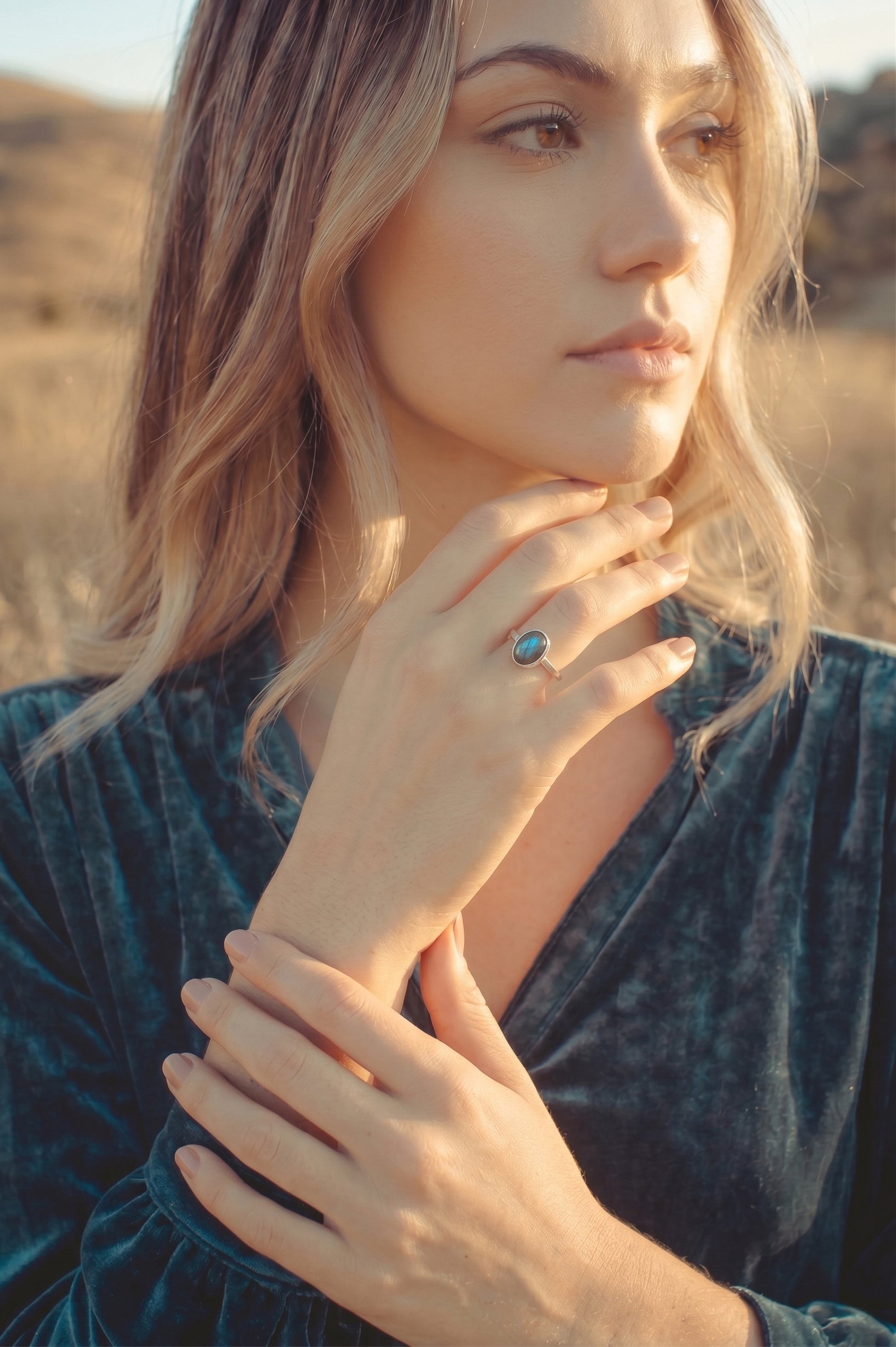 Model wearing a dainty oval labradorite ring in sterling silver on her finger. Lifestyle photography showcasing a blue flash gemstone ring paired with a teal velvet dress in golden hour sunlight.