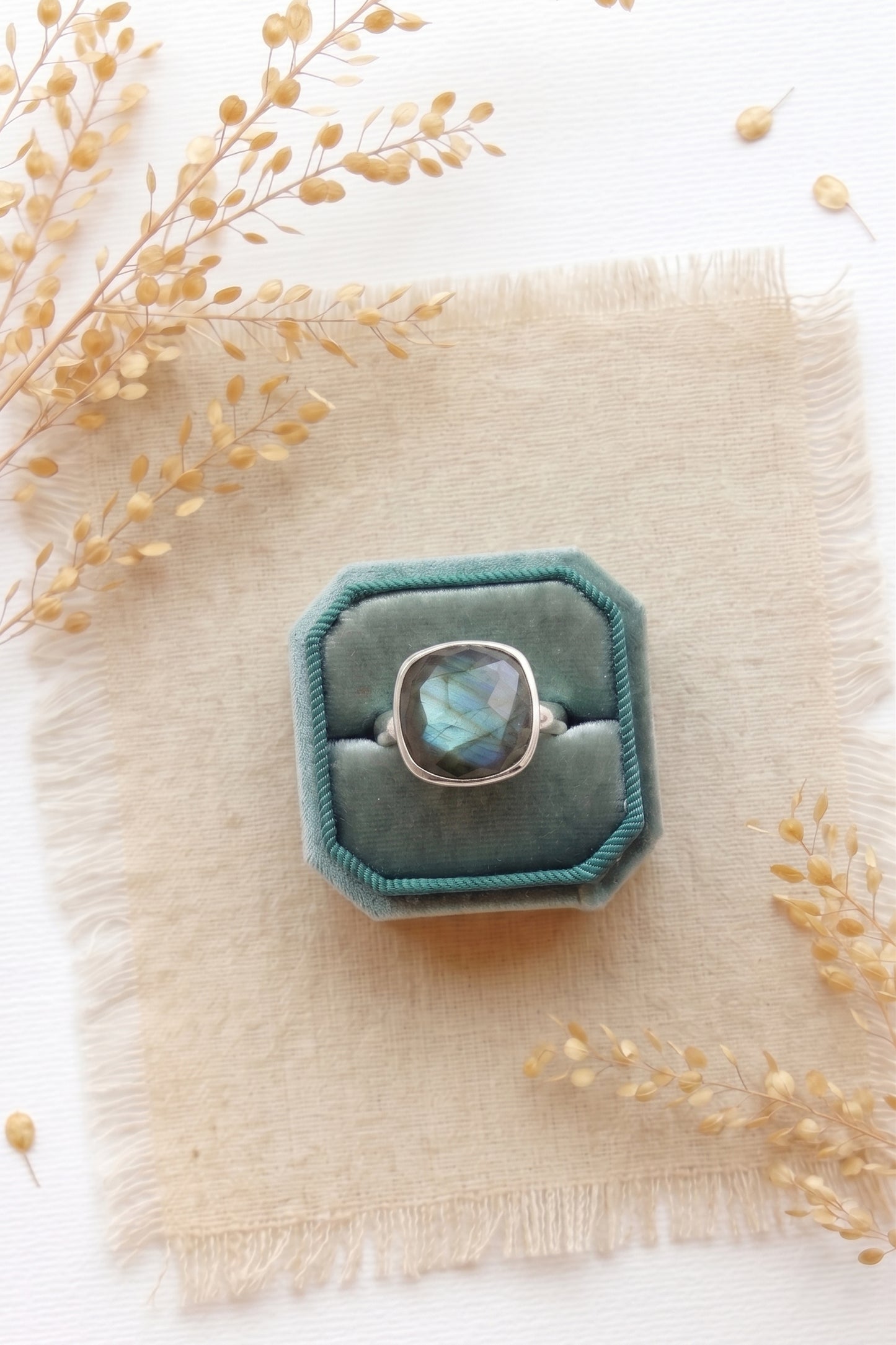 Portrait of a blonde woman wearing a deep teal velvet dress standing in a sunlit field of golden grass. She holds one hand up to her chin, elegantly showcasing a prominent, flashing blue cushion-cut labradorite silver cocktail ring on her middle finger.